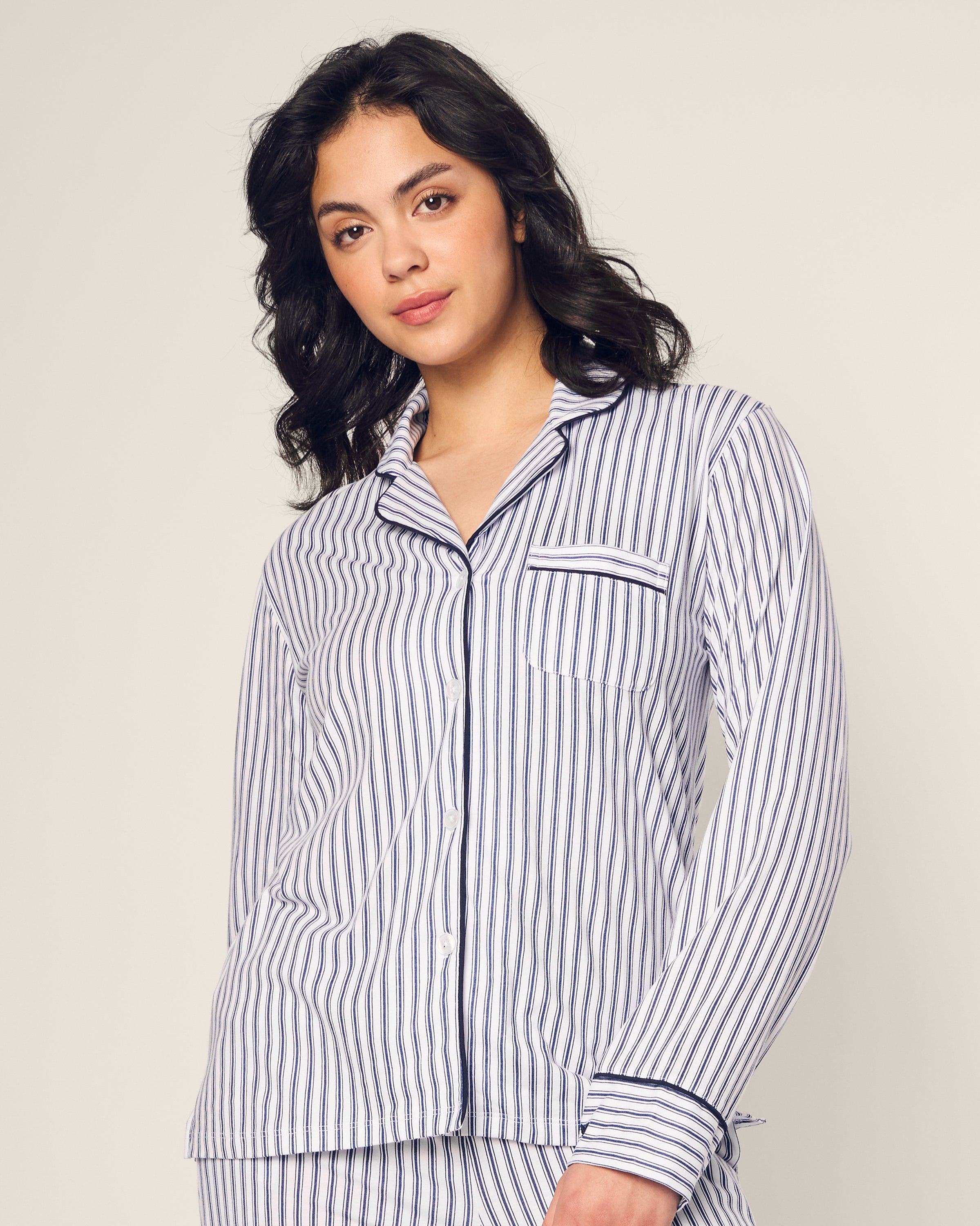 The individual with long, curly hair is wearing the Womens Pima Pajama Set in Navy French Ticking by Petite Plume, crafted from luxurious Peruvian Pima cotton, featuring a striped design with a button-up top and front pocket.