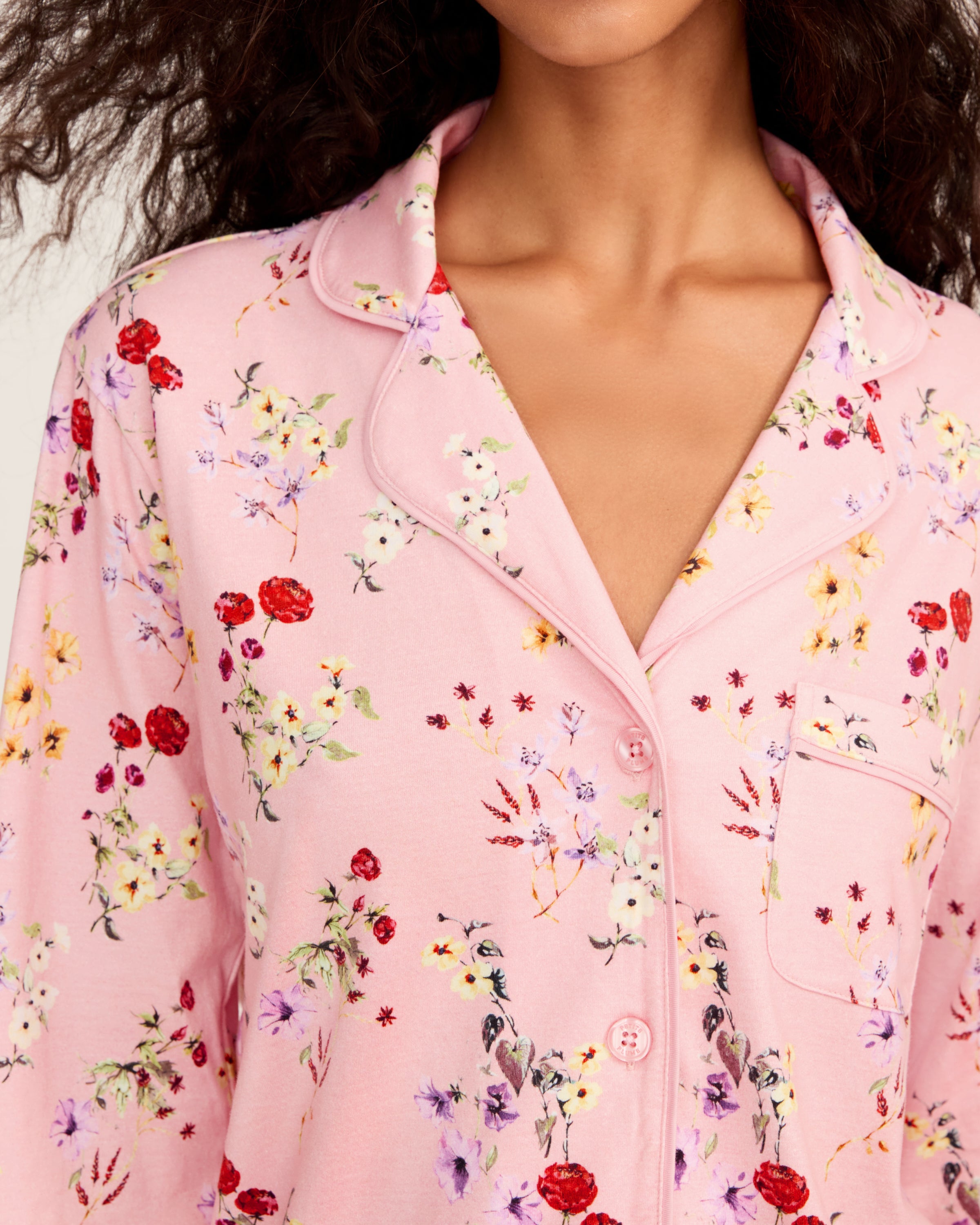 The Women's Pima Pajama Set in Brilliant Botanical Blush features a pink button-up long-sleeve top and matching pants, both adorned with a vibrant floral print in red, white, and purple.