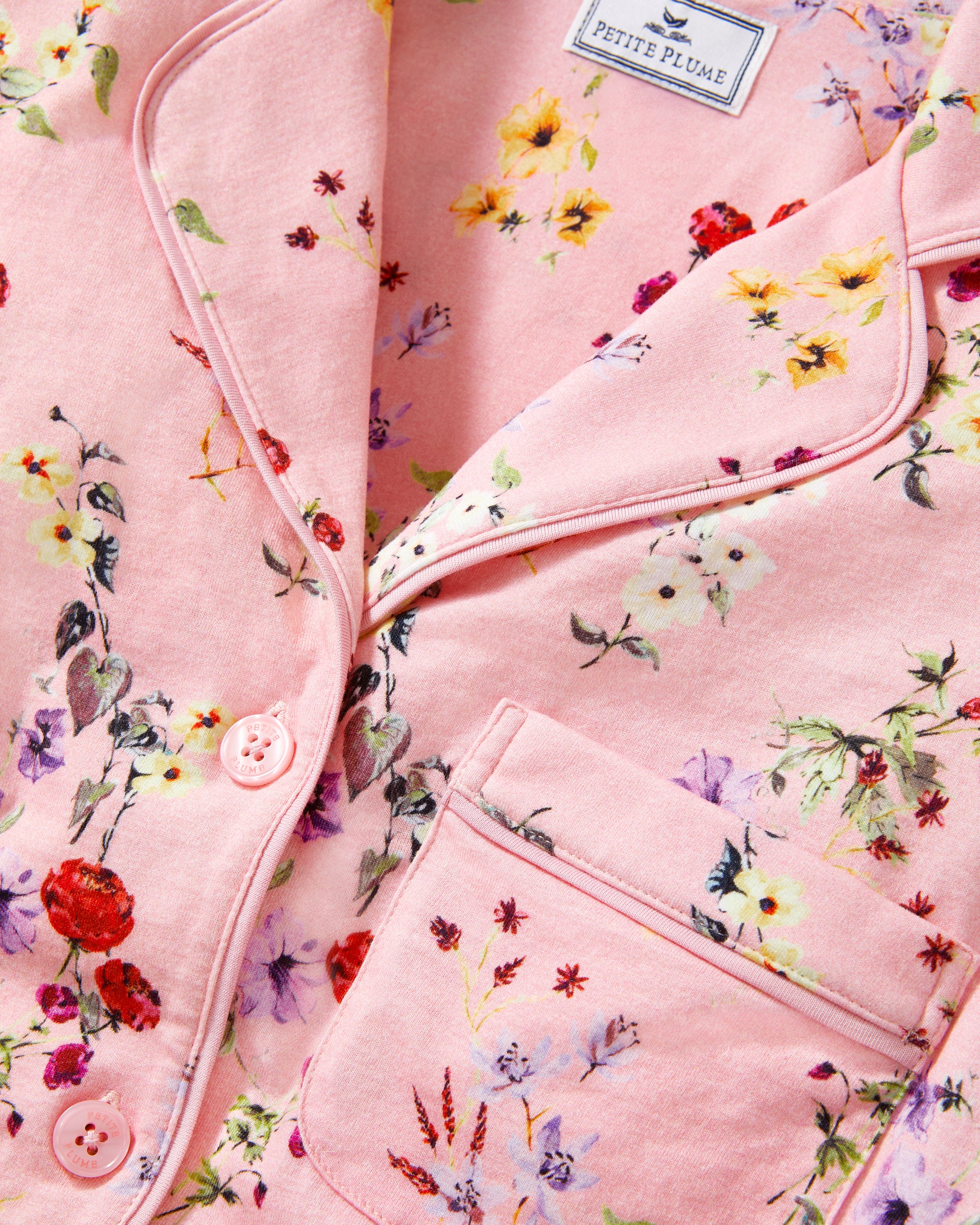 The Women's Pima Pajama Set in Brilliant Botanical Blush features a pink button-up long-sleeve top and matching pants, both adorned with a vibrant floral print in red, white, and purple.