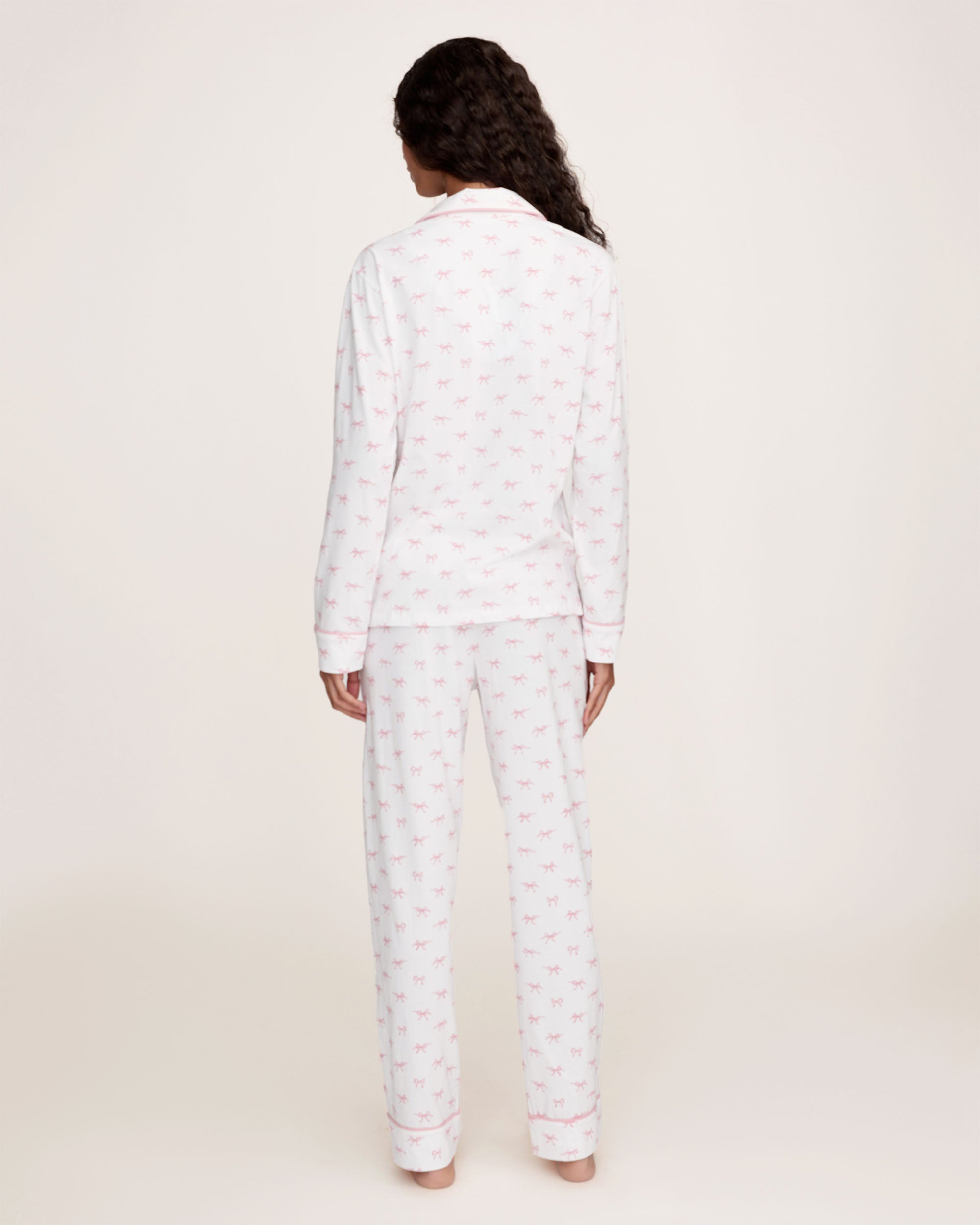 The Women's Pima Pajama Set in Blushing Bows features a long-sleeve button-up top and pants in white fabric with a small pink bow pattern and pink piping accents.