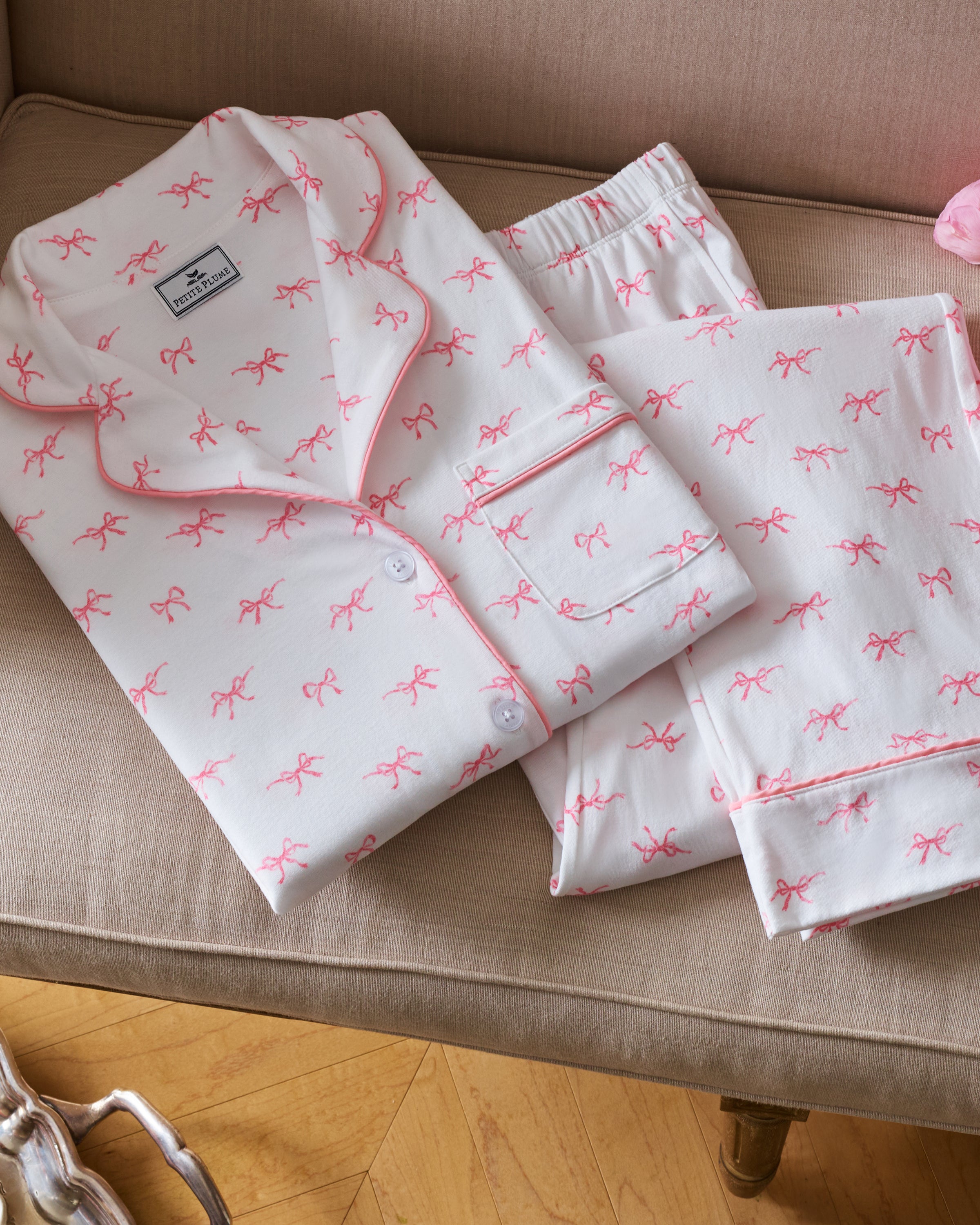 The Women's Pima Pajama Set in Blushing Bows features a long-sleeve button-up top and pants in white fabric with a small pink bow pattern and pink piping accents.