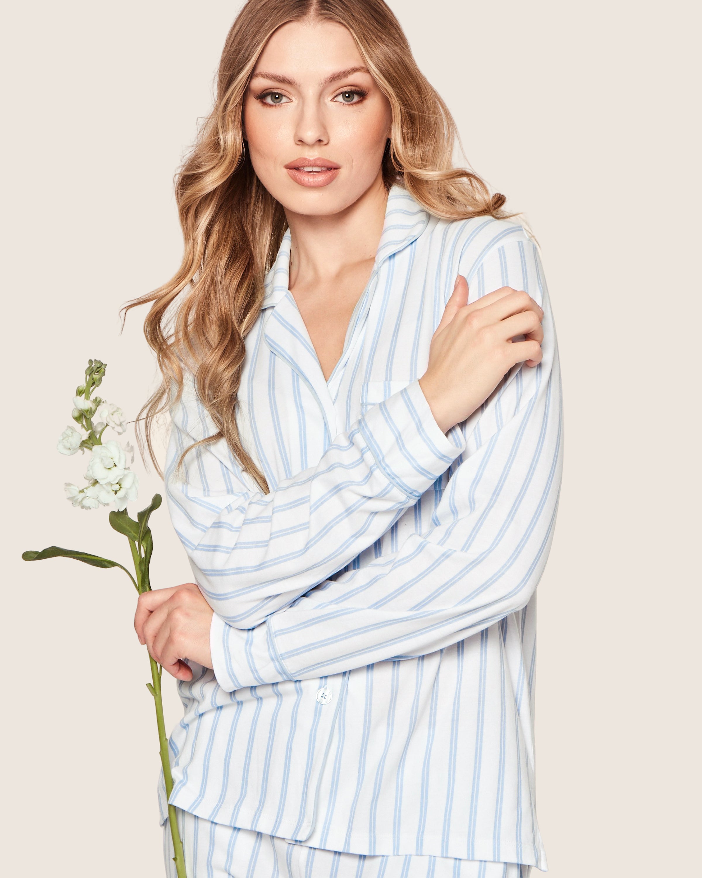A woman with long, wavy blonde hair wears Petite Plumes Womens Pima Pajama Set in Periwinkle and White Stripe, holding a white flower and gazing at the camera against a beige background.