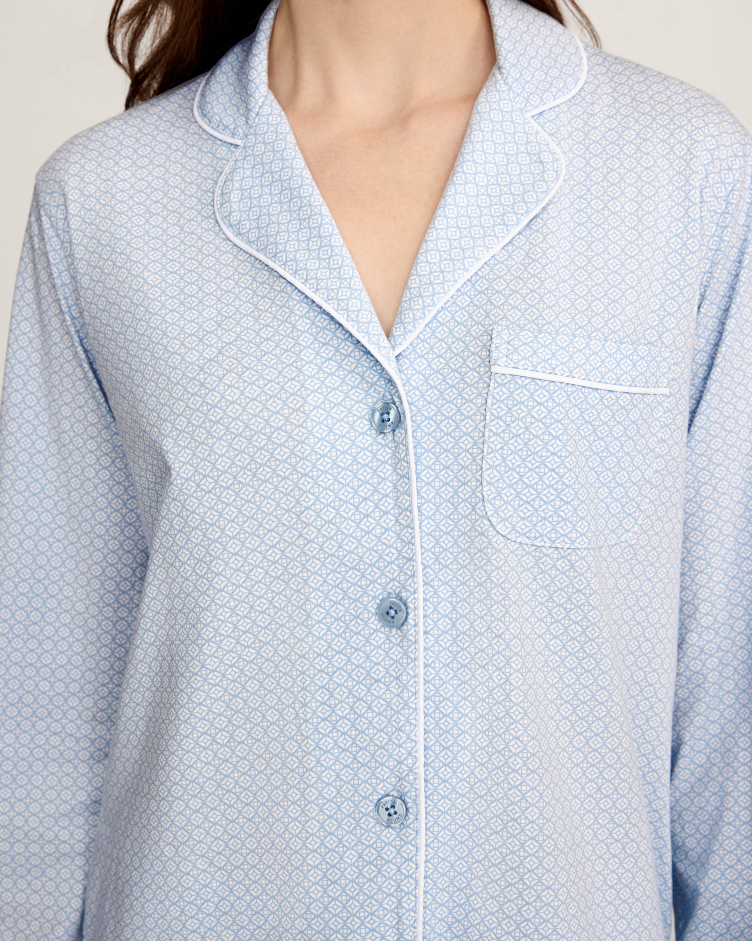 A woman with light brown hair and a neutral expression stands barefoot against a plain background, modeling the Women's Lightweight Pima Pajama Set in Periwinkle Geo—featuring long sleeves, button-up top with collar, and matching pants.