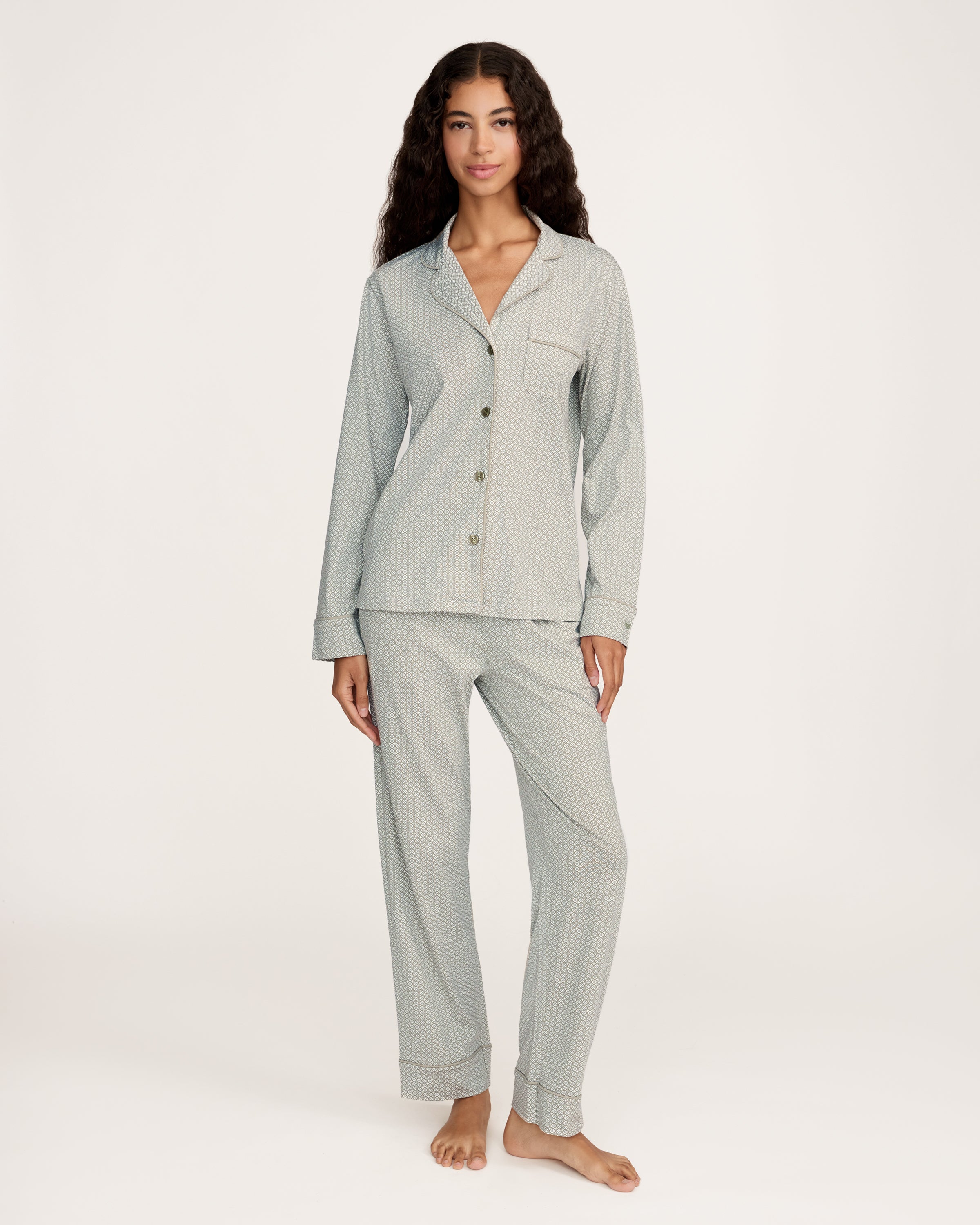 A woman with long curly hair stands barefoot against a white background, wearing the Women's Lightweight Pima Pajama Set in Sage Geo—a light gray button-up long-sleeve top and matching pants with a small geometric pattern.