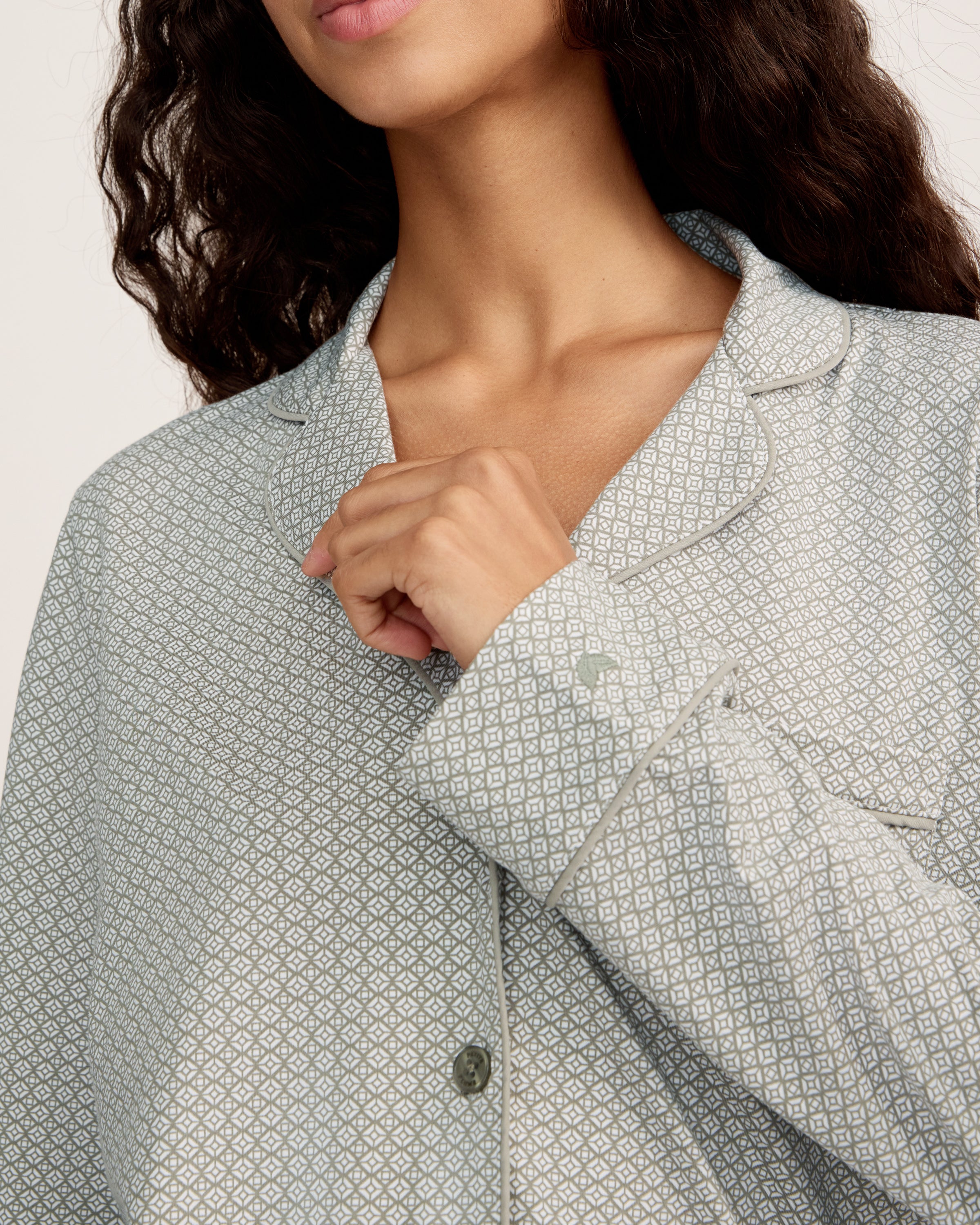 A woman with long curly hair stands barefoot against a white background, wearing the Women's Lightweight Pima Pajama Set in Sage Geo—a light gray button-up long-sleeve top and matching pants with a small geometric pattern.