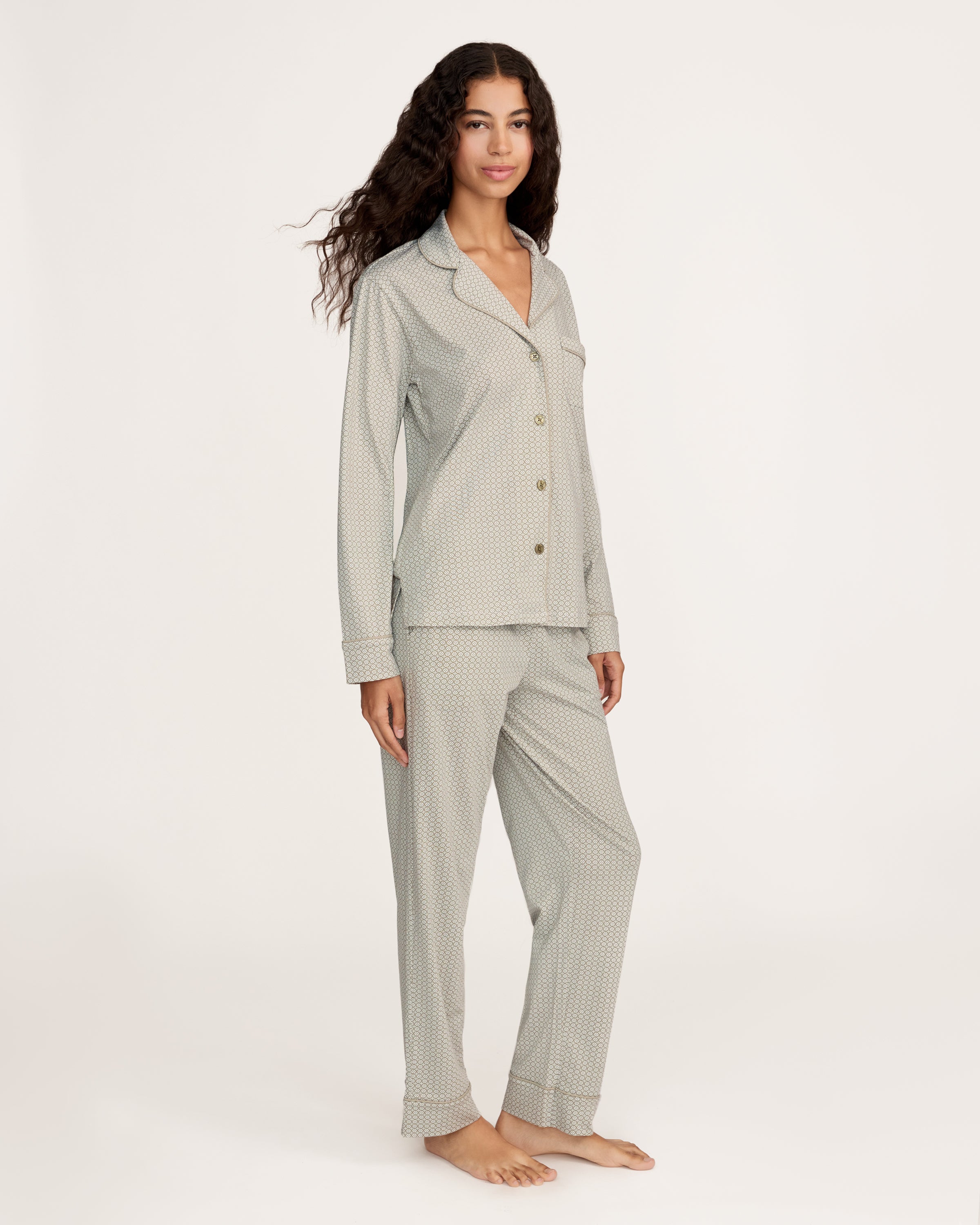 A woman with long curly hair stands barefoot against a white background, wearing the Women's Lightweight Pima Pajama Set in Sage Geo—a light gray button-up long-sleeve top and matching pants with a small geometric pattern.
