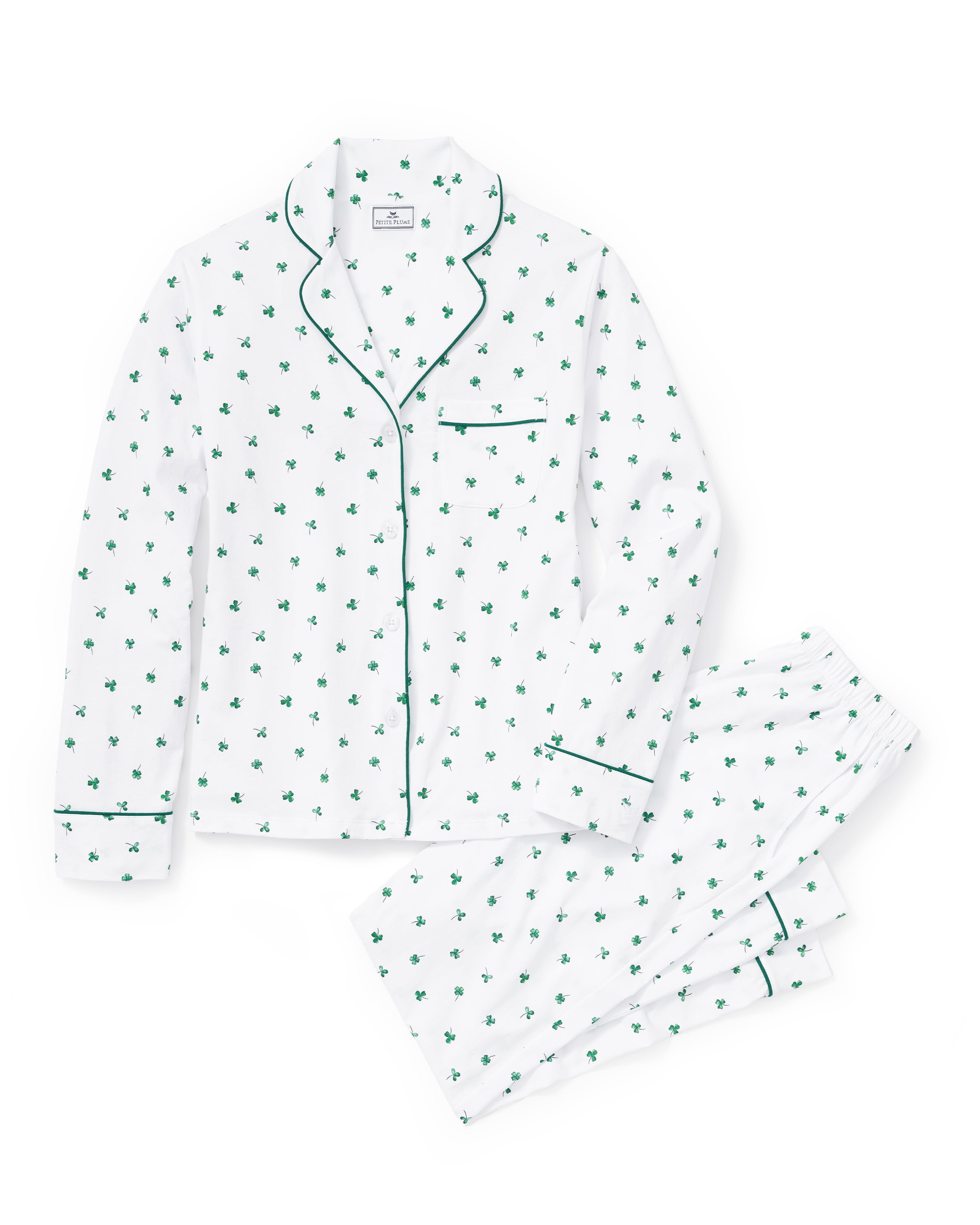 A woman with long dark hair stands barefoot on a white background, wearing the Women's Pima Pajama Set in Shamrocks—white pajama top and pants featuring a green shamrock print and green piping. She has a neutral expression.