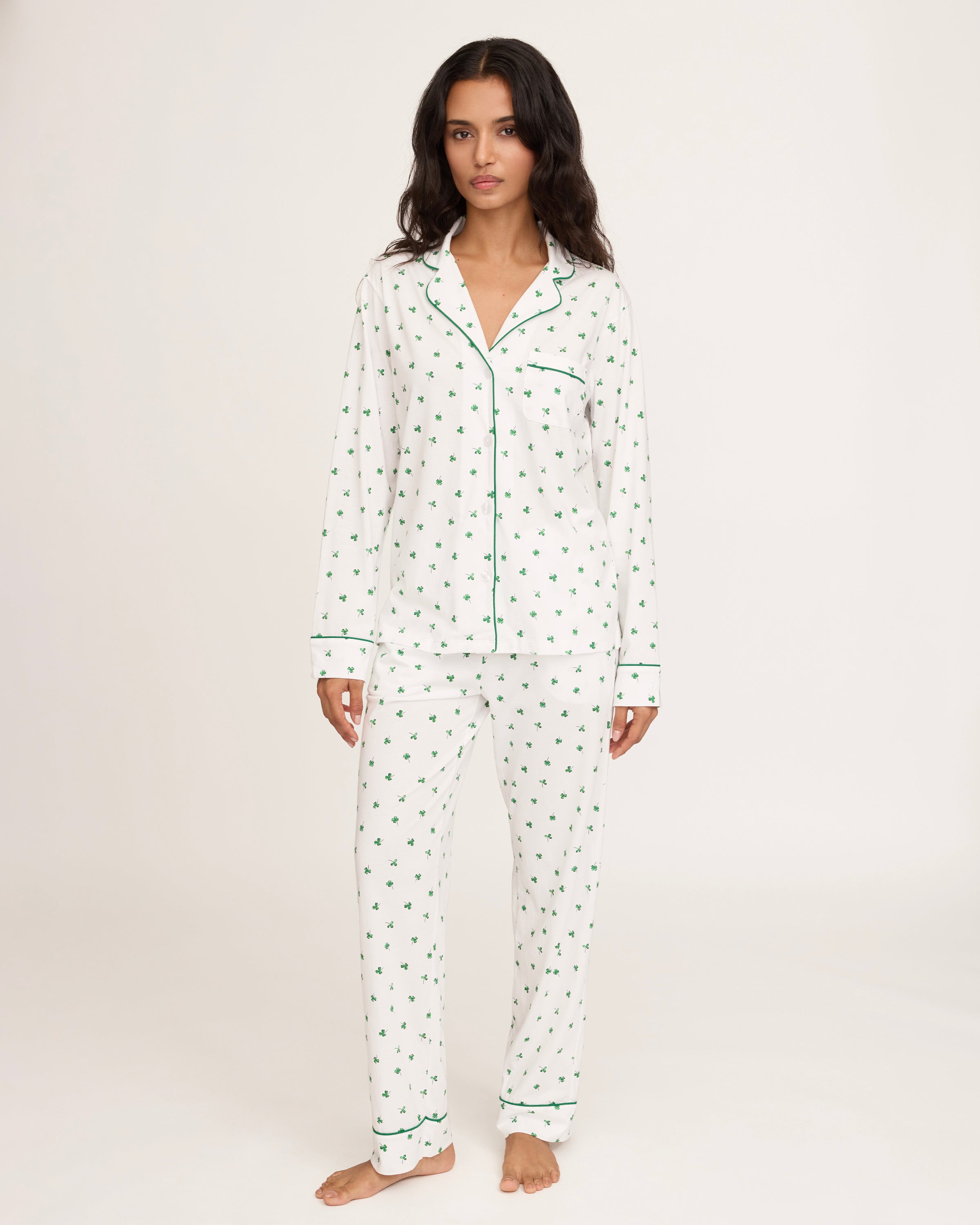 A woman with long dark hair stands barefoot on a white background, wearing the Women's Pima Pajama Set in Shamrocks—white pajama top and pants featuring a green shamrock print and green piping. She has a neutral expression.