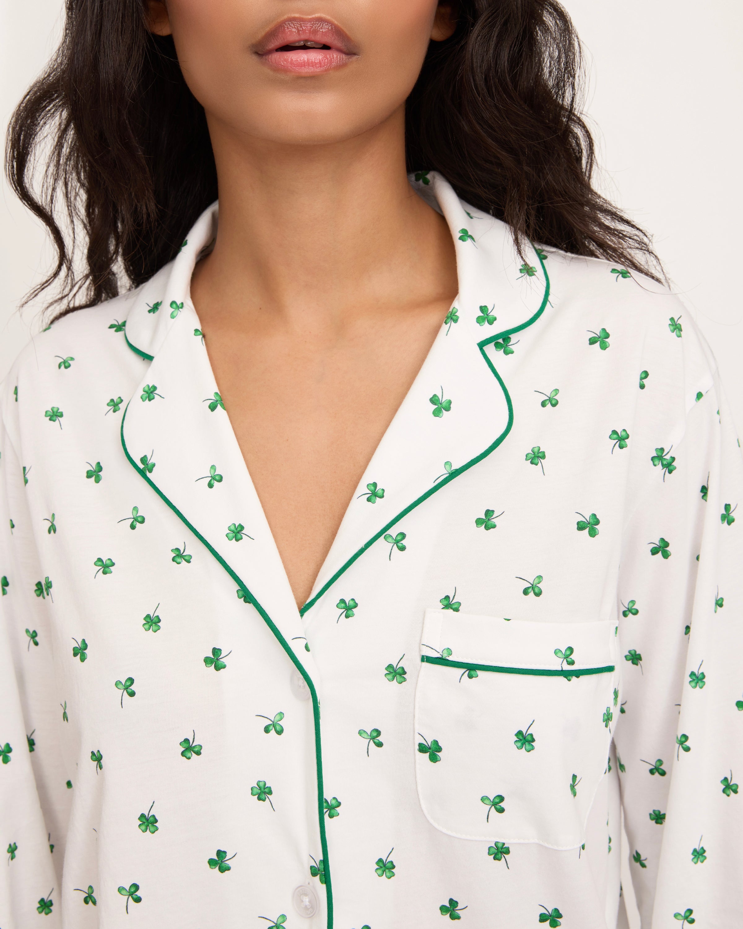 A woman with long dark hair stands barefoot on a white background, wearing the Women's Pima Pajama Set in Shamrocks—white pajama top and pants featuring a green shamrock print and green piping. She has a neutral expression.