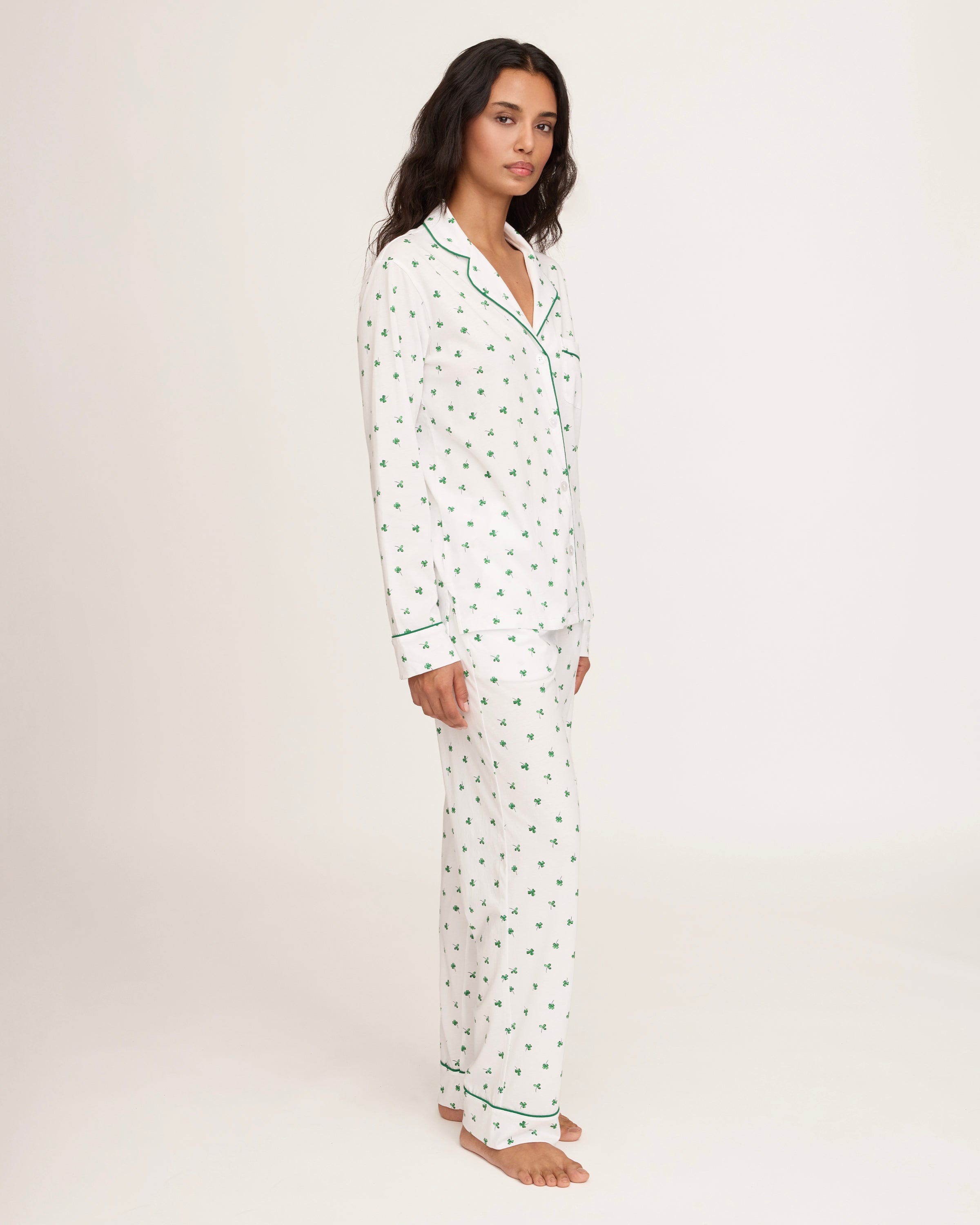 A woman with long dark hair stands barefoot on a white background, wearing the Women's Pima Pajama Set in Shamrocks—white pajama top and pants featuring a green shamrock print and green piping. She has a neutral expression.