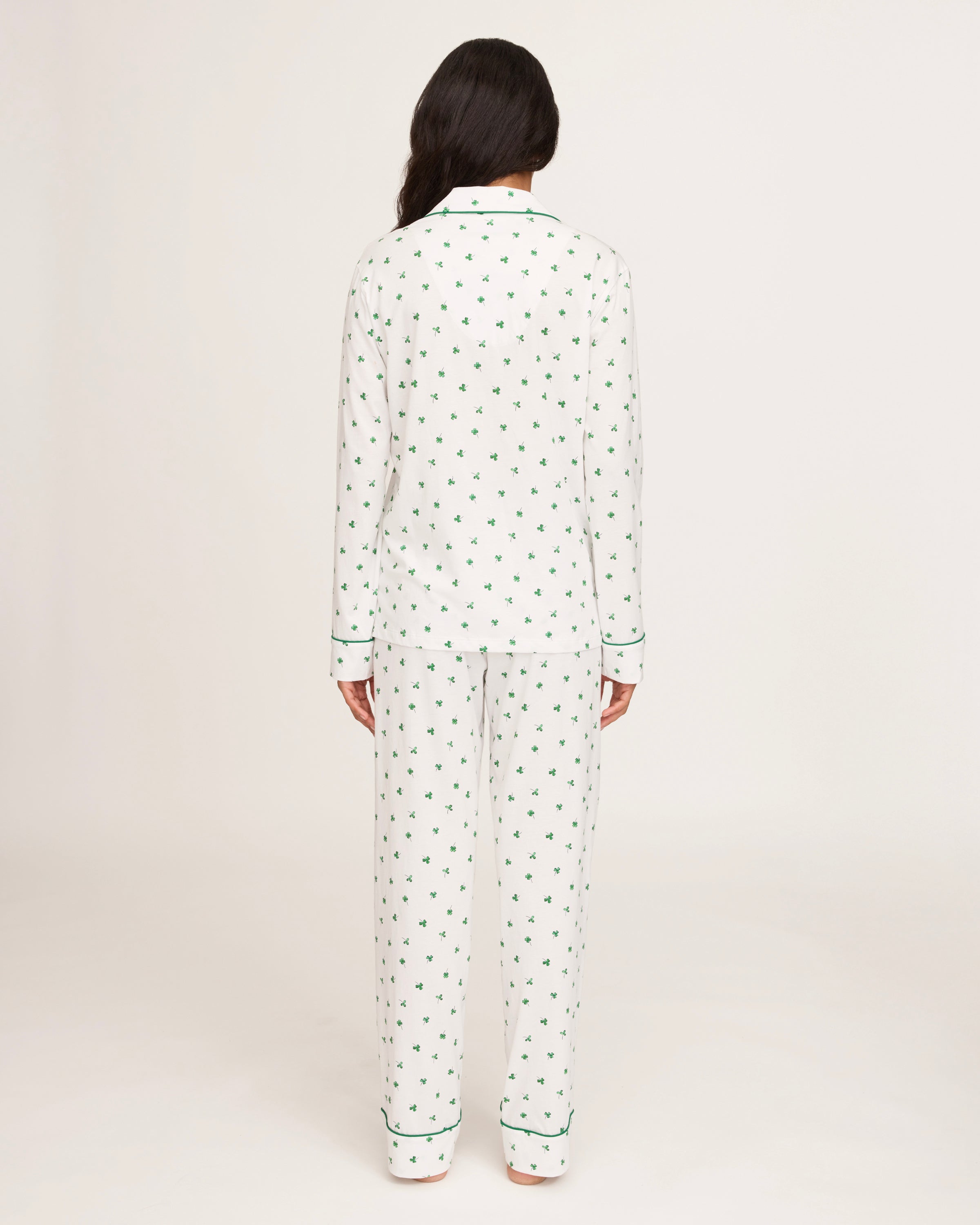 A woman with long dark hair stands barefoot on a white background, wearing the Women's Pima Pajama Set in Shamrocks—white pajama top and pants featuring a green shamrock print and green piping. She has a neutral expression.