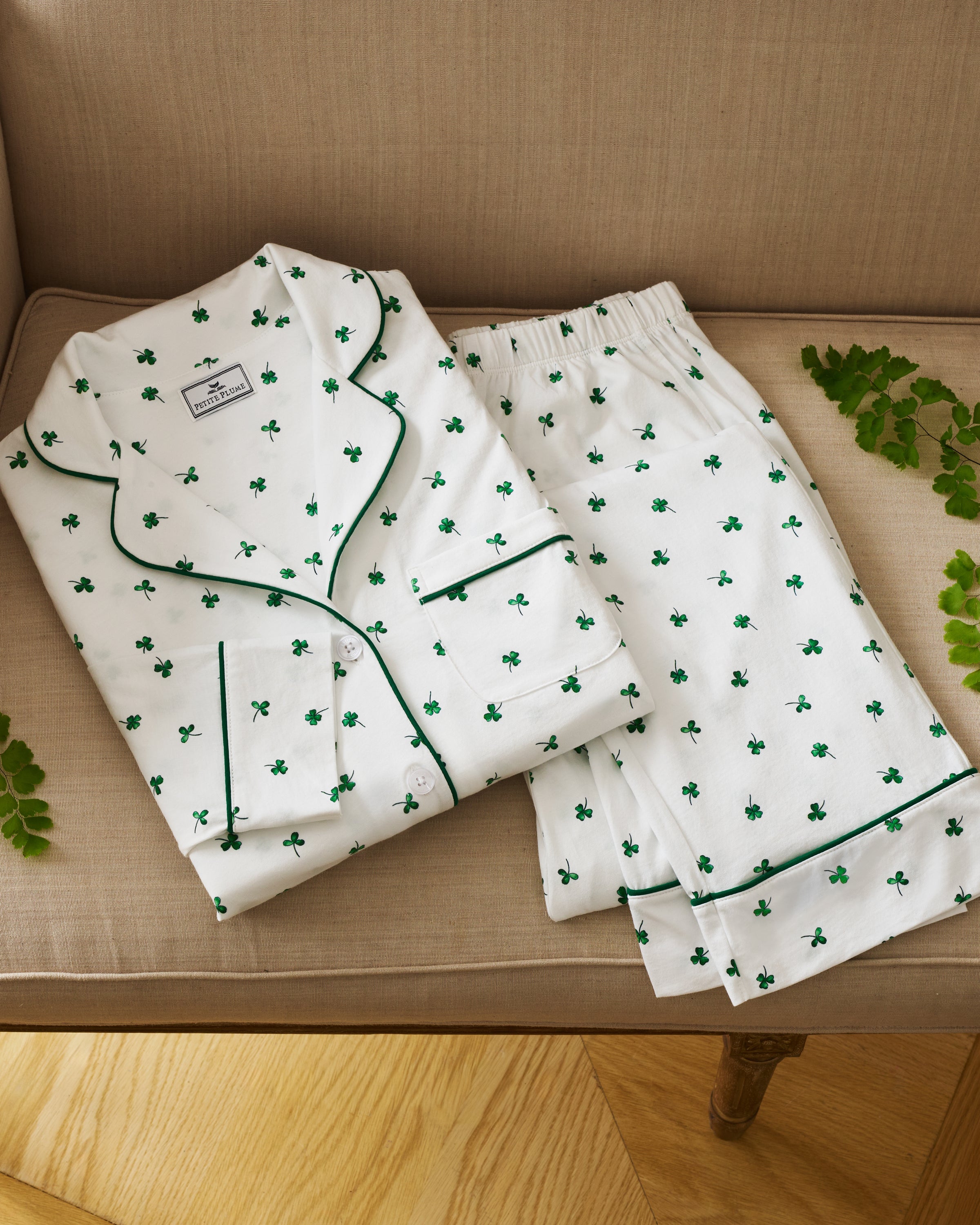 A woman with long dark hair stands barefoot on a white background, wearing the Women's Pima Pajama Set in Shamrocks—white pajama top and pants featuring a green shamrock print and green piping. She has a neutral expression.
