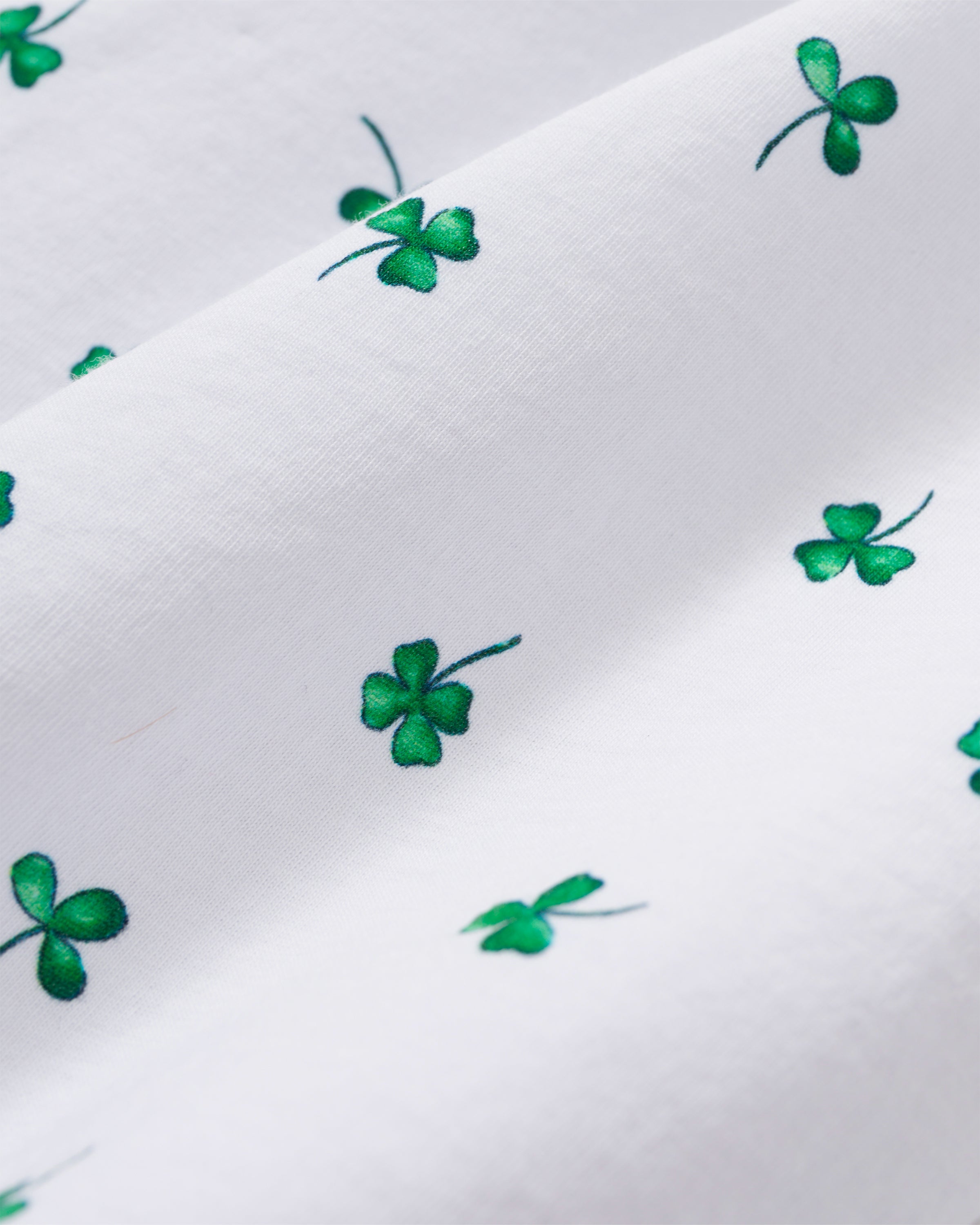 A woman with long dark hair stands barefoot on a white background, wearing the Women's Pima Pajama Set in Shamrocks—white pajama top and pants featuring a green shamrock print and green piping. She has a neutral expression.