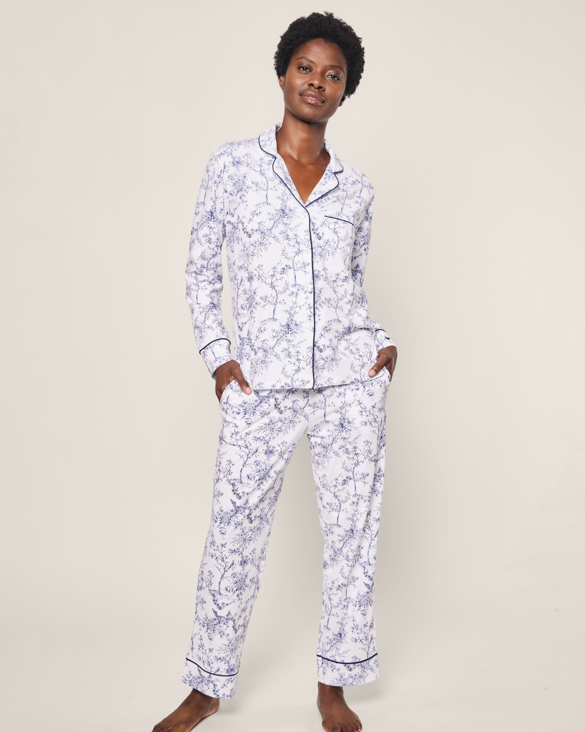 A person wearing Petite Plume’s Women’s Pima Pajama Set in Timeless Toile stands against a plain background. The luxury sleepwear features a button-up top and long pants, both accented with dark piping. They have their hands in the pockets and a relaxed posture.