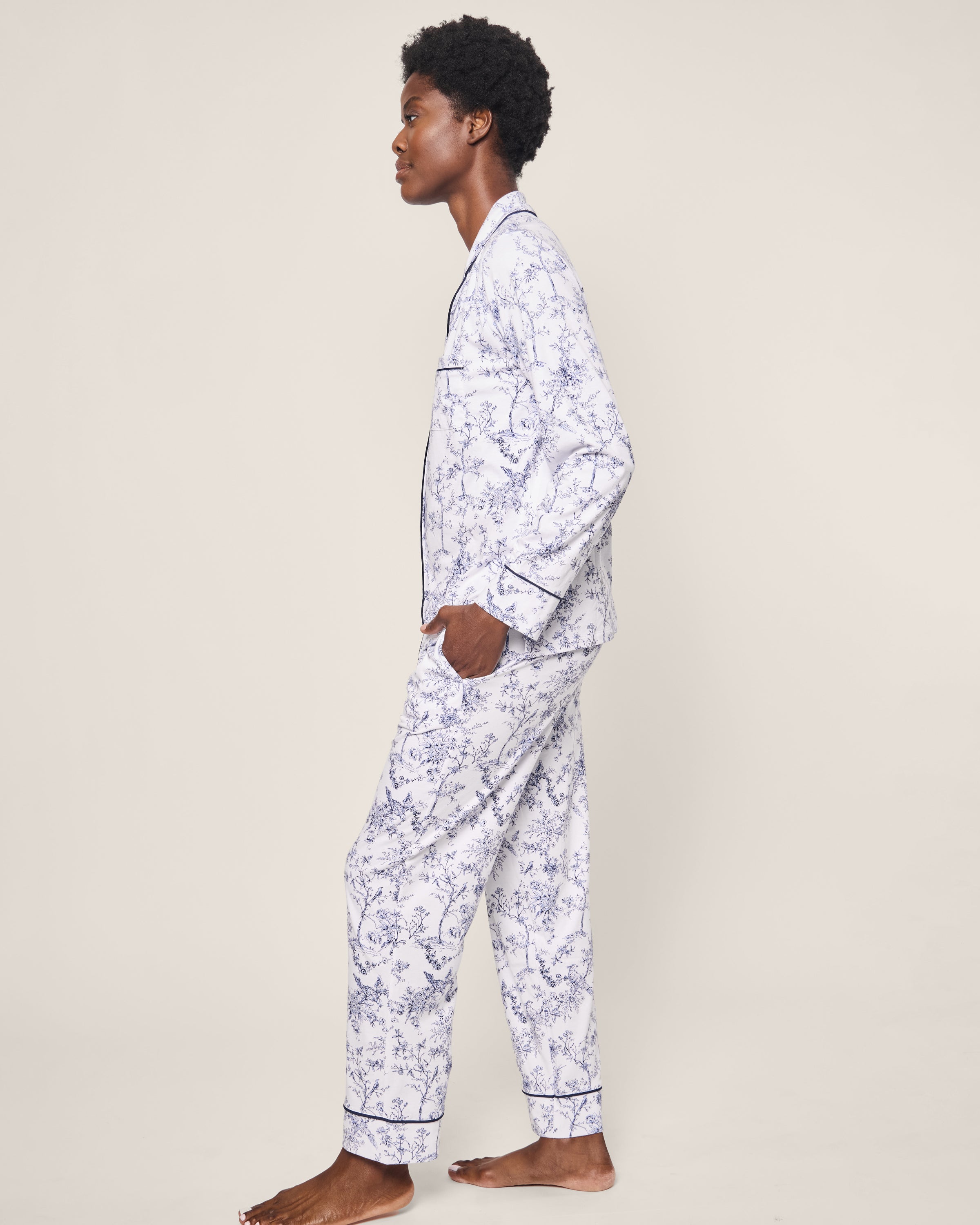 A person stands barefoot in profile, wearing the luxurious Womens Pima Pajama Set in Timeless Toile, featuring a light blue and white floral pattern with dark trim. The plain, light-colored background highlights the botanical elegance of this classic Petite Plume sleepwear ensemble.