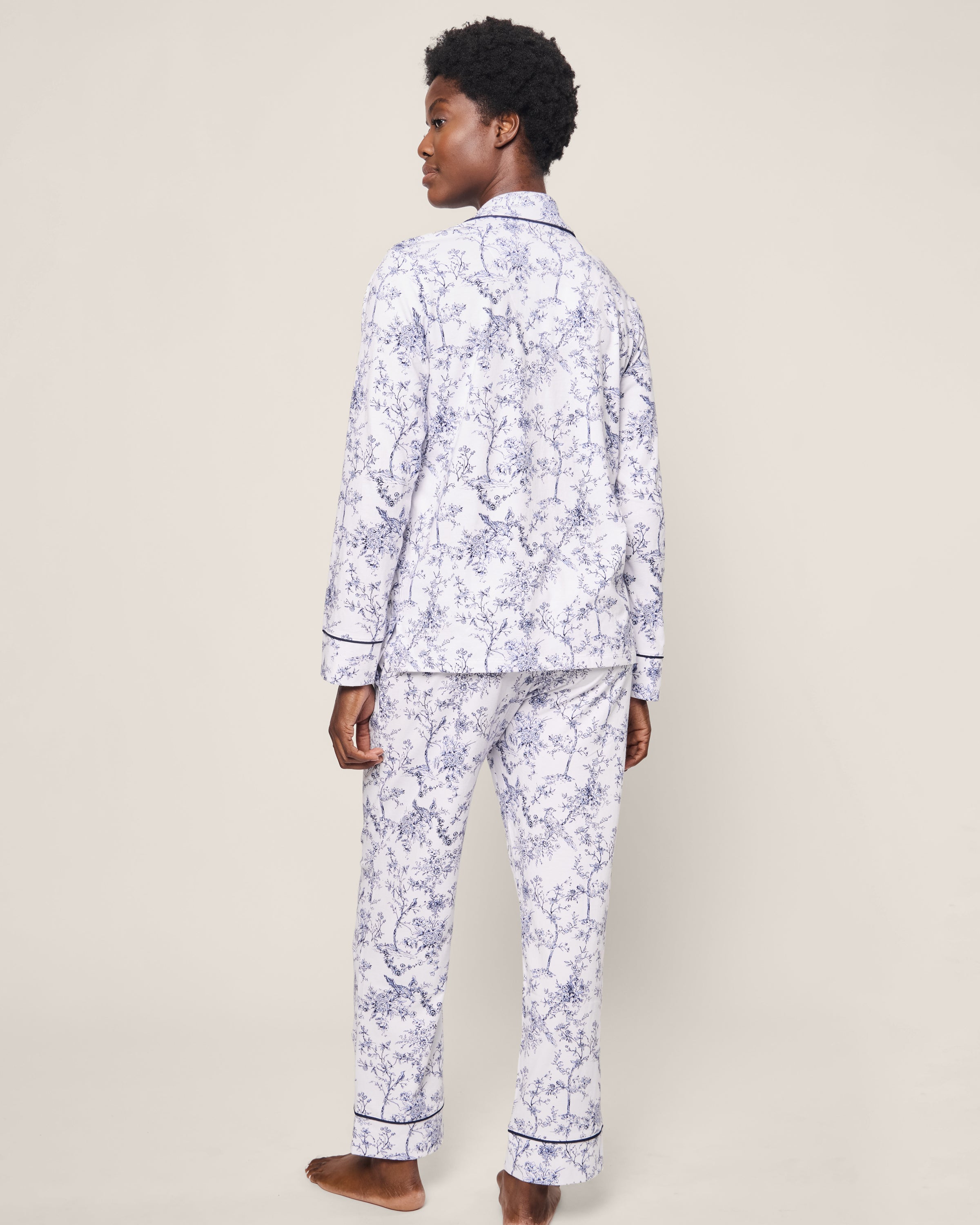 A person models the Petite Plume Womens Pima Pajama Set in Timeless Toile, showcasing its luxurious floral-patterned white design with blue accents. The long-sleeve and pant set is highlighted as they stand barefoot against a plain background, glancing over their shoulder.