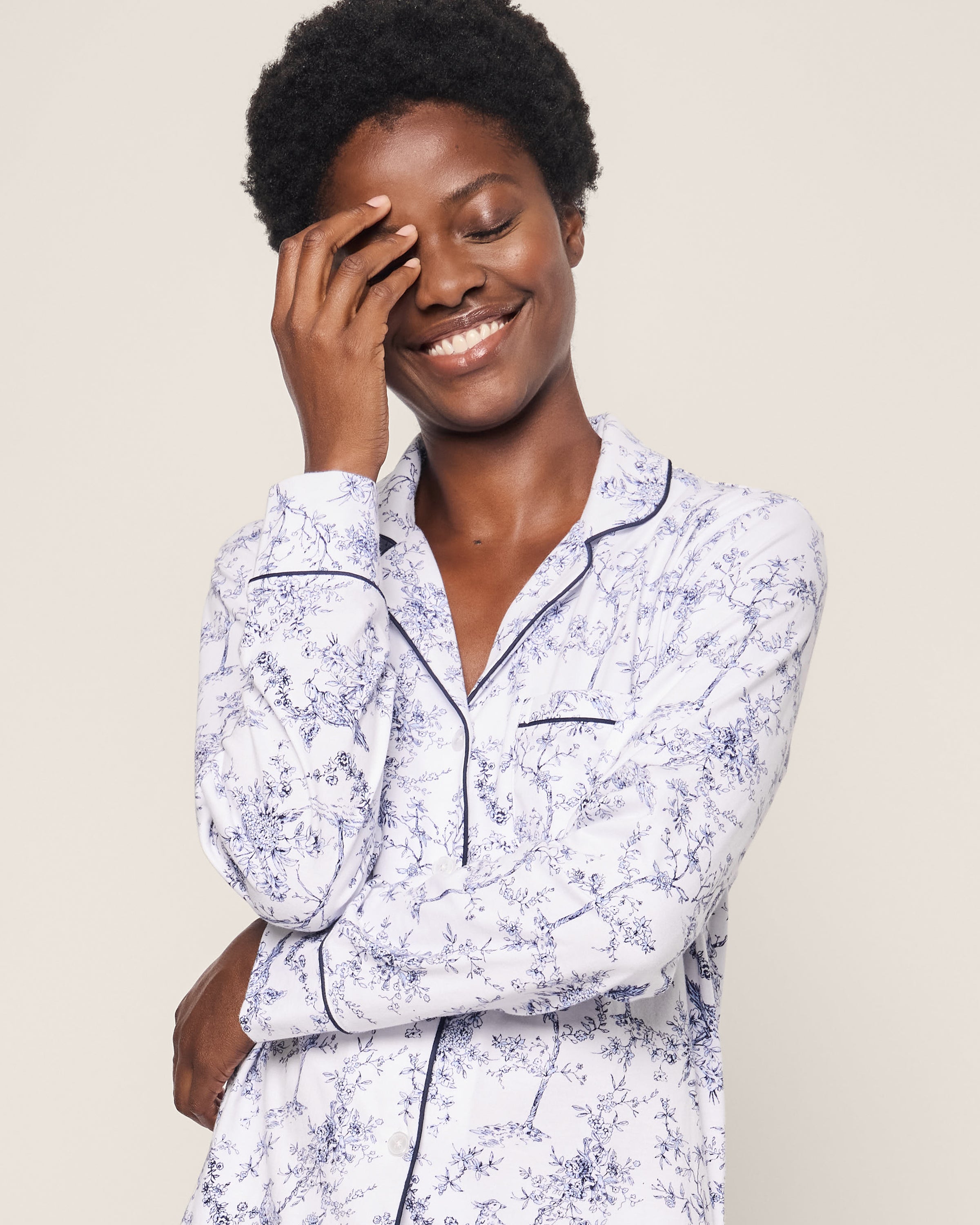 A smiling person with short, curly hair wears the Petite Plume Womens Pima Pajama Set in Timeless Toile, featuring a long-sleeved floral-patterned top. One hand rests on their forehead, the other arm is crossed, all against a neutral background.