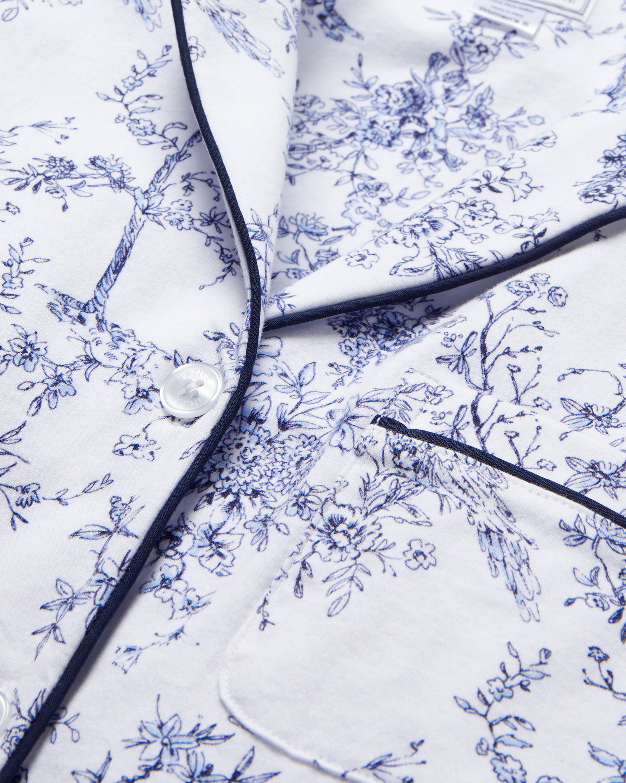 The Womens Pima Pajama Set in Timeless Toile by Petite Plume features a white fabric with intricate blue botanical patterns, dark piping, buttons, and a pocket. It is perfect for those who appreciate classic floral designs.
