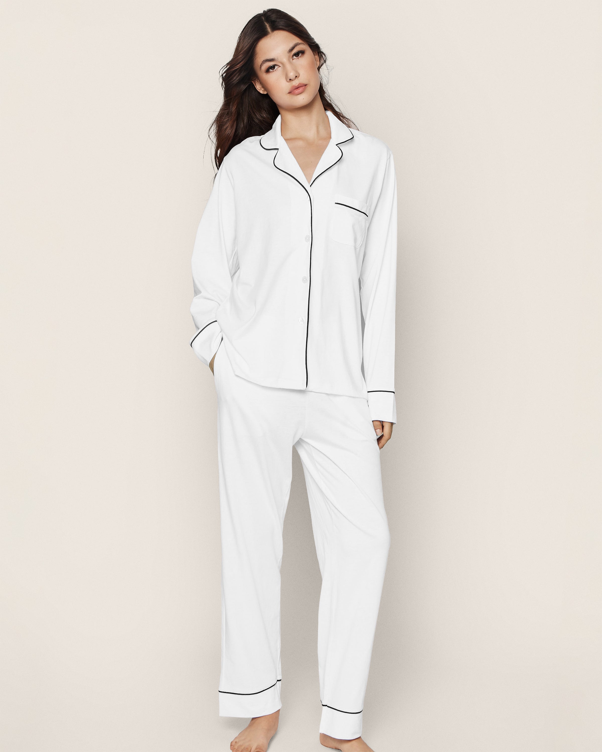 A person with long dark hair, barefoot against a plain background, wears Petite Plumes luxurious Womens Pima Pajama Set in White with Navy Piping, featuring a long-sleeve button-up shirt and pants.