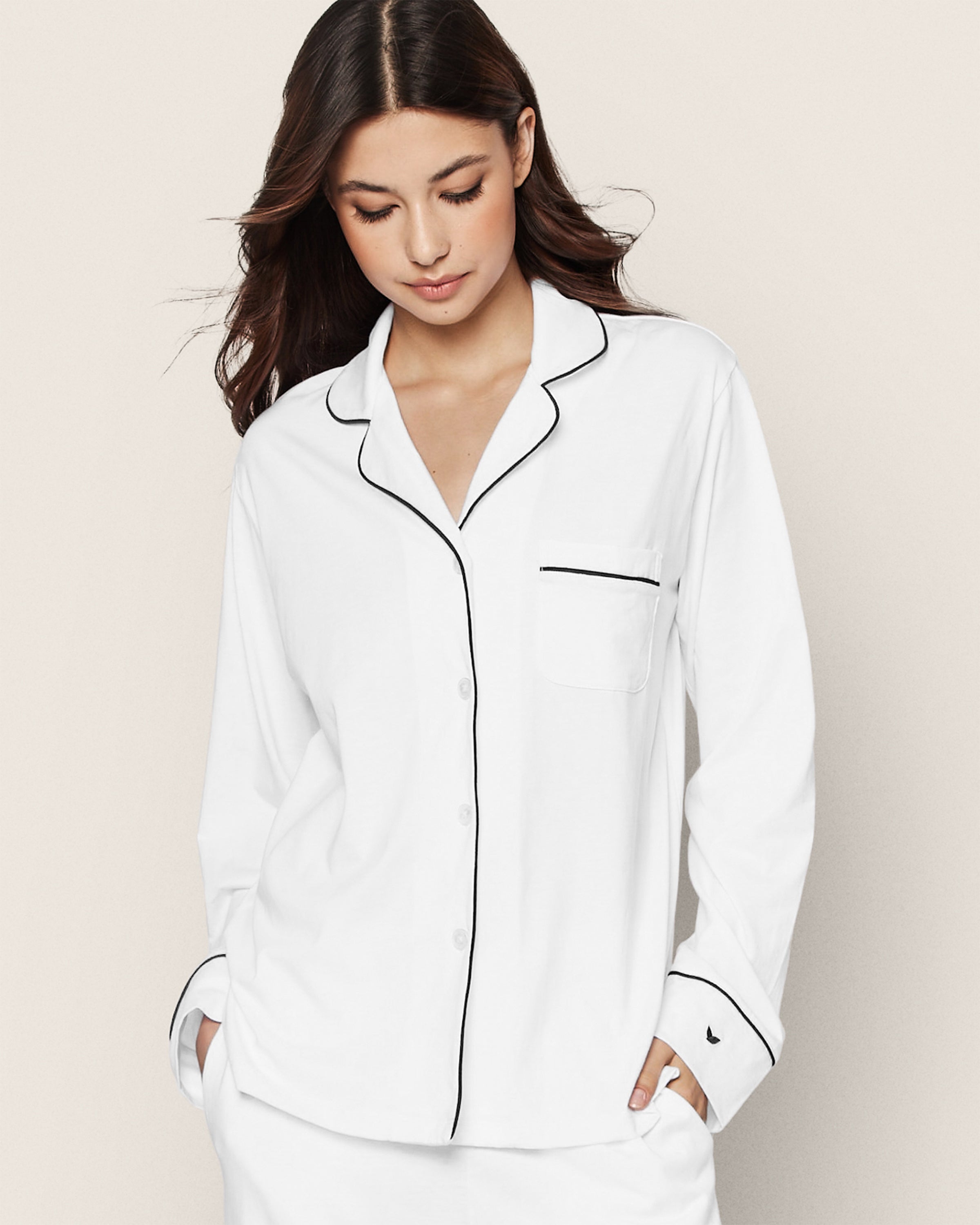 A woman with long, dark hair wears the Petite Plume Womens Pima Pajama Set in White with Navy Piping, featuring a white top and matching pants crafted from luxurious Peruvian Pima cotton. She gazes down with her hands tucked into the pockets.