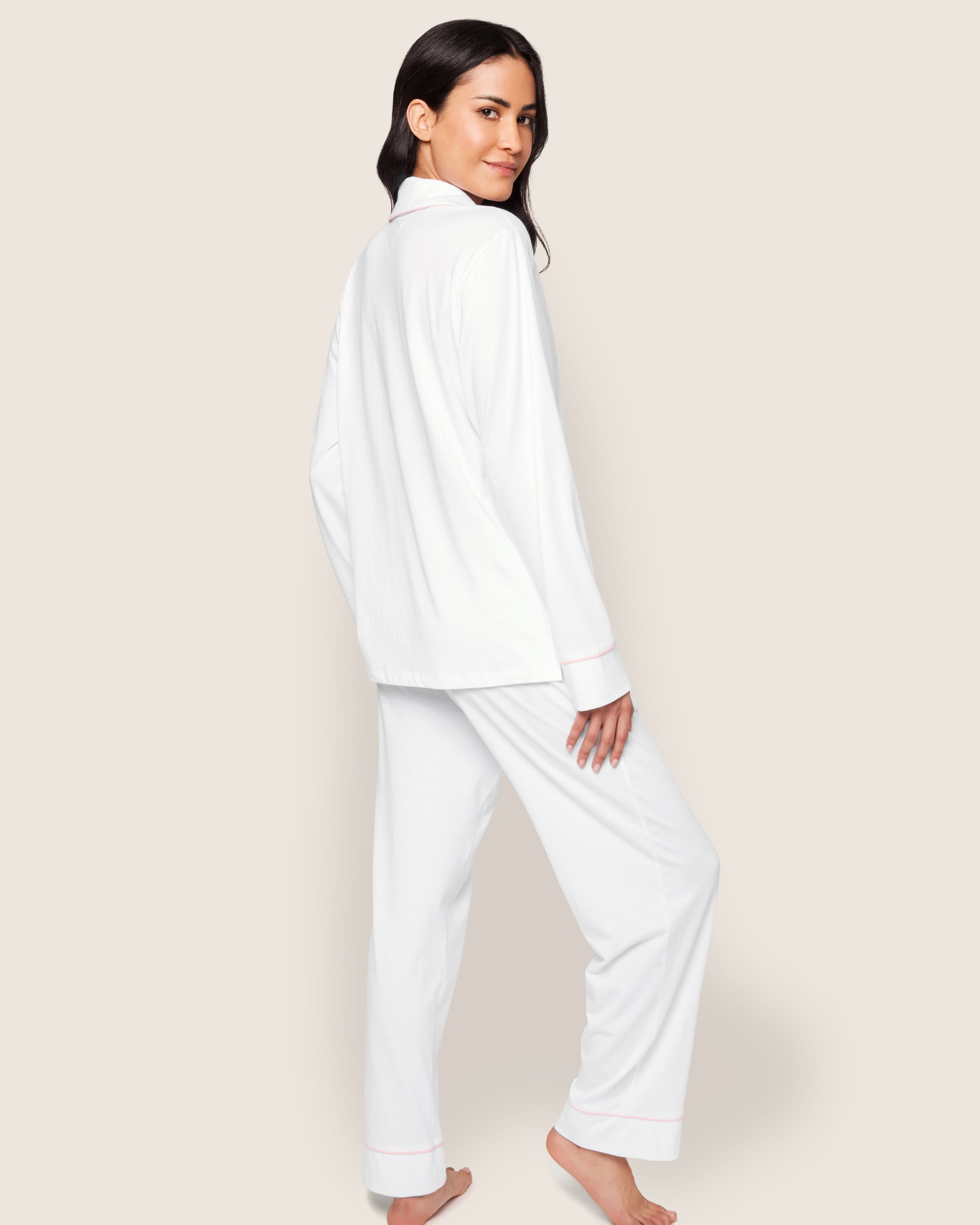 A person with long dark hair stands barefoot, turning their head towards the camera, wearing the elegant Womens Pima Pajama Set in White with Pink Piping by Petite Plume. The plain light background enhances the luxurious Peruvian Pima cotton ensemble.