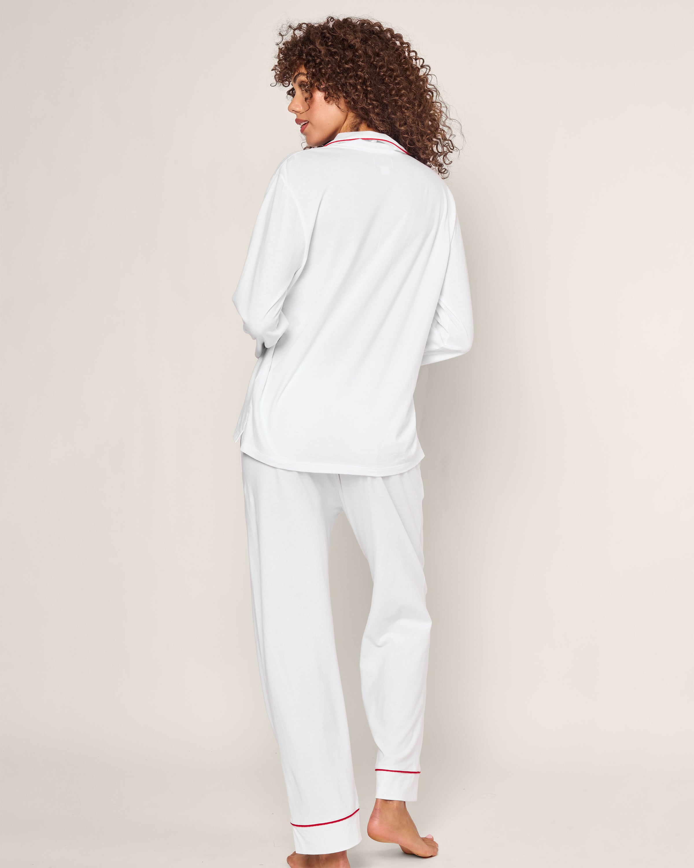 A curly-haired person, barefoot on a light background, models the timeless silhouette of Petite Plumes Womens White Pima Pajama Set with Red Piping. Crafted from luxurious Peruvian Pima cotton, they turn away to highlight the outfits side and back.