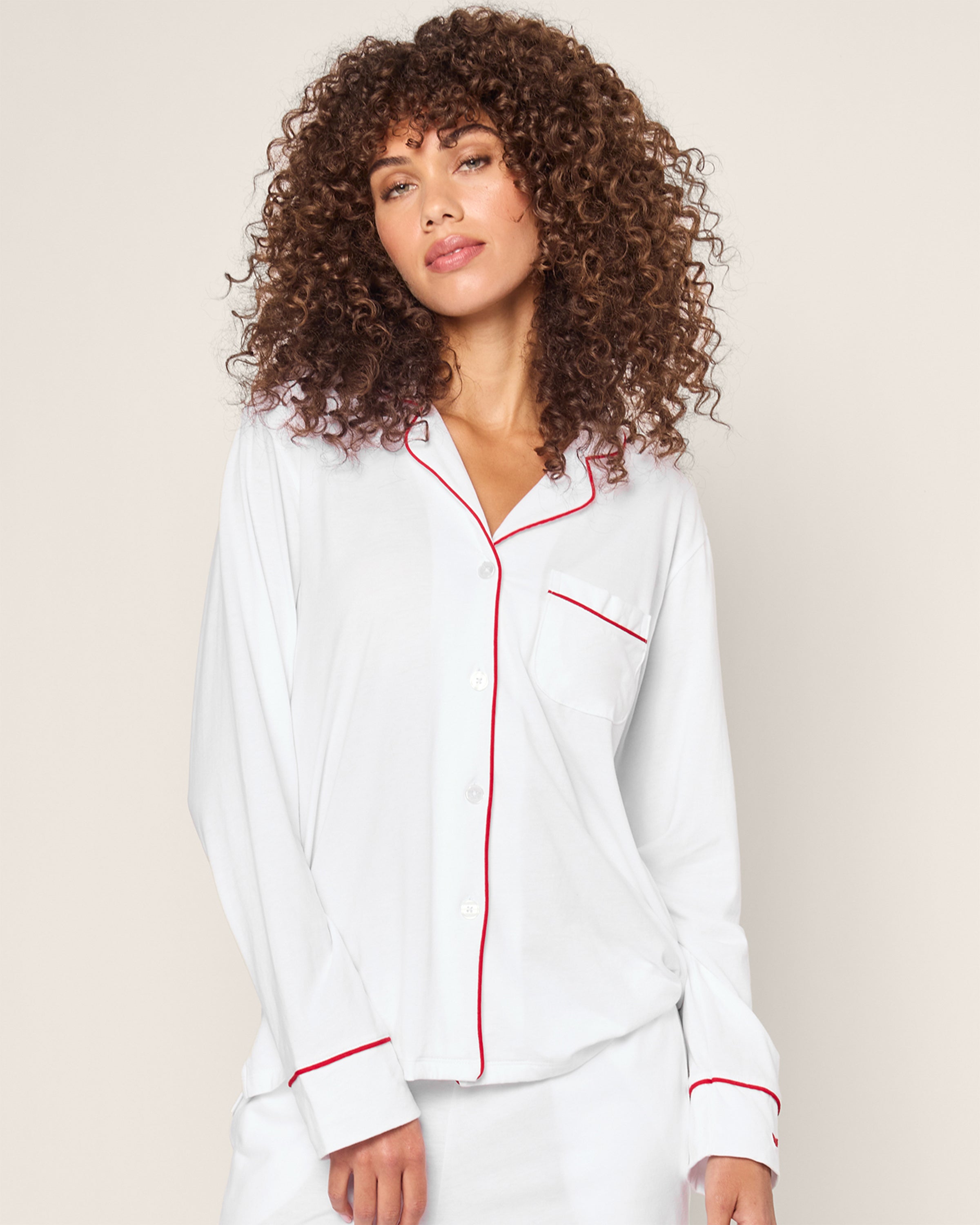 A person with curly hair wears Petite Plumes Womens White Pima Pajama Set with Red Piping, highlighting its timeless style. Made from luxurious Peruvian Pima cotton, it boasts a left pocket and front buttons against a plain, light background.