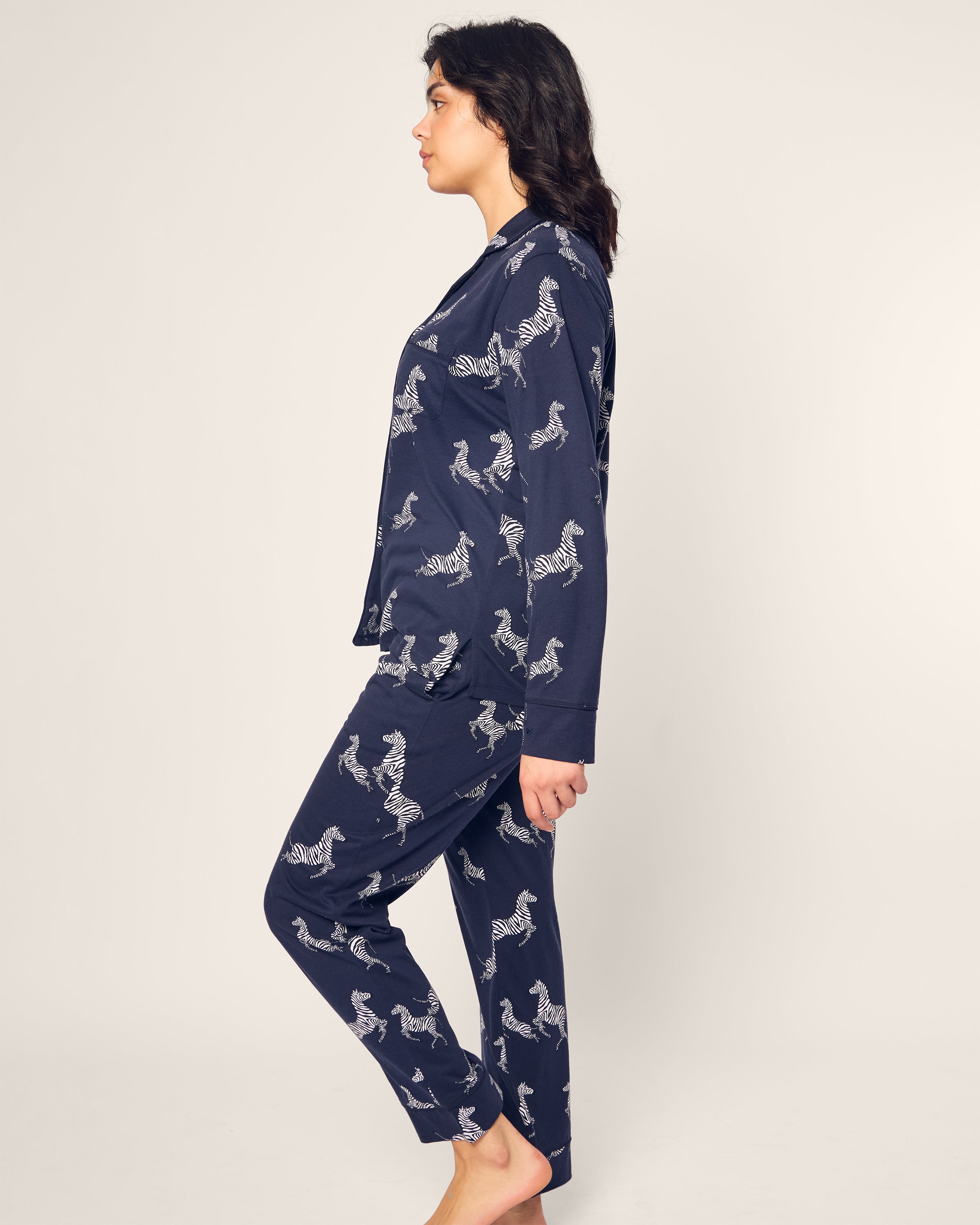 A person in profile stands barefoot against a plain backdrop, draped in the Womens Pima Pajama Set in Zèbre de Nuit: navy blue luxury sleepwear adorned with elegant white zebra prints, crafted from soft Pima cotton.