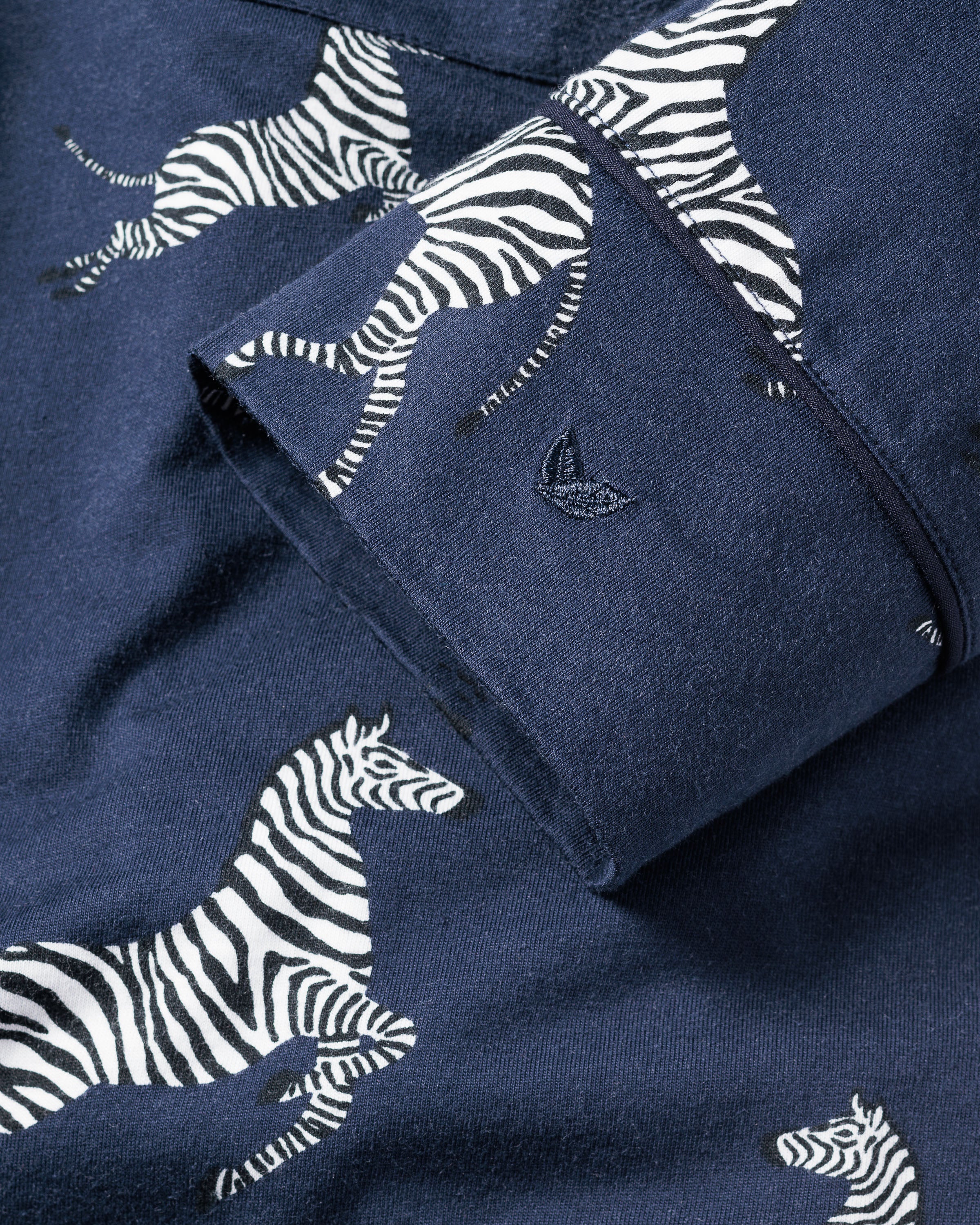 The Womens Pima Pajama Set in Zèbre de Nuit by Petite Plume showcases navy blue fabric with a white and black zebra pattern. Made from premium Peruvian Pima cotton for comfort, it features a cuff adorned with a subtle sailboat logo embroidery.