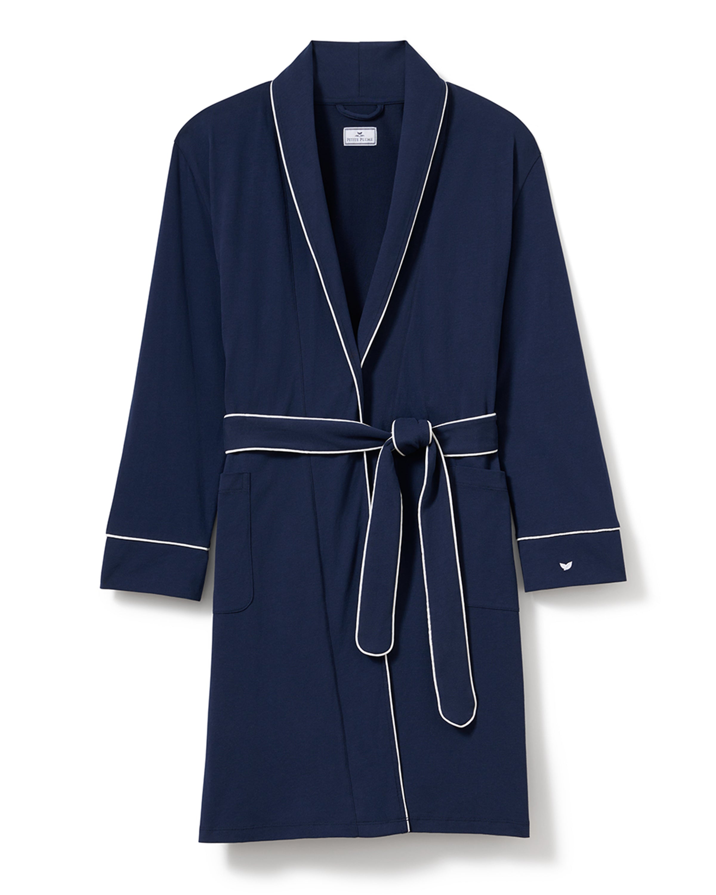 A woman in the Petite Plume Womens Pima Maternity Robe in Navy with white piping and a waist belt stands against a light background. Made of soft pima cotton, she has long dark hair, looks down gently smiling, and rests one hand on her abdomen.