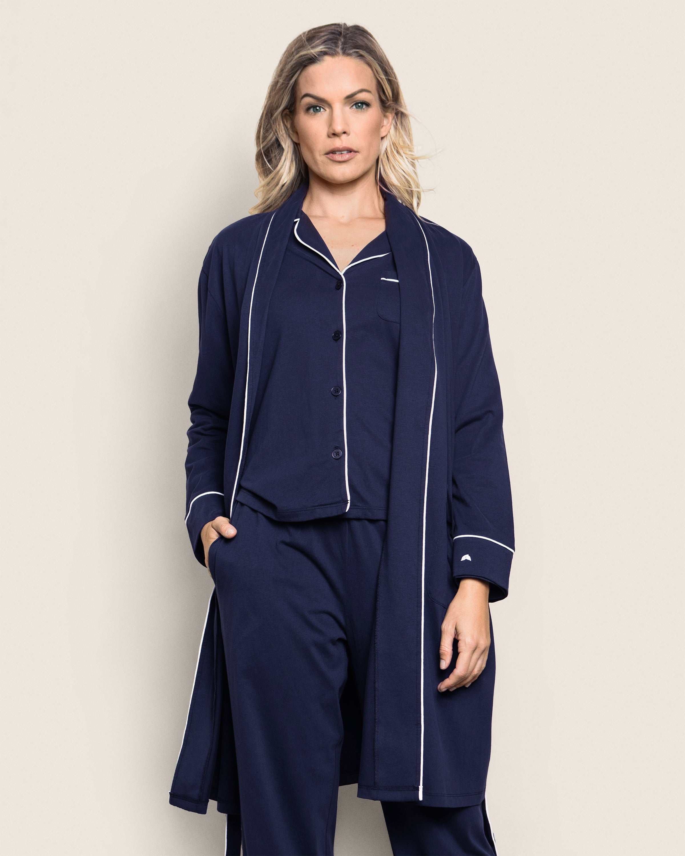 A person with long hair stands confidently in dark blue luxury pajamas made from yarn-dyed Peruvian Pima cotton, highlighted with white piping. They have the Womens Pima Robe in Navy by Petite Plume draped over their shoulders, hands in pockets, set against a light beige background.