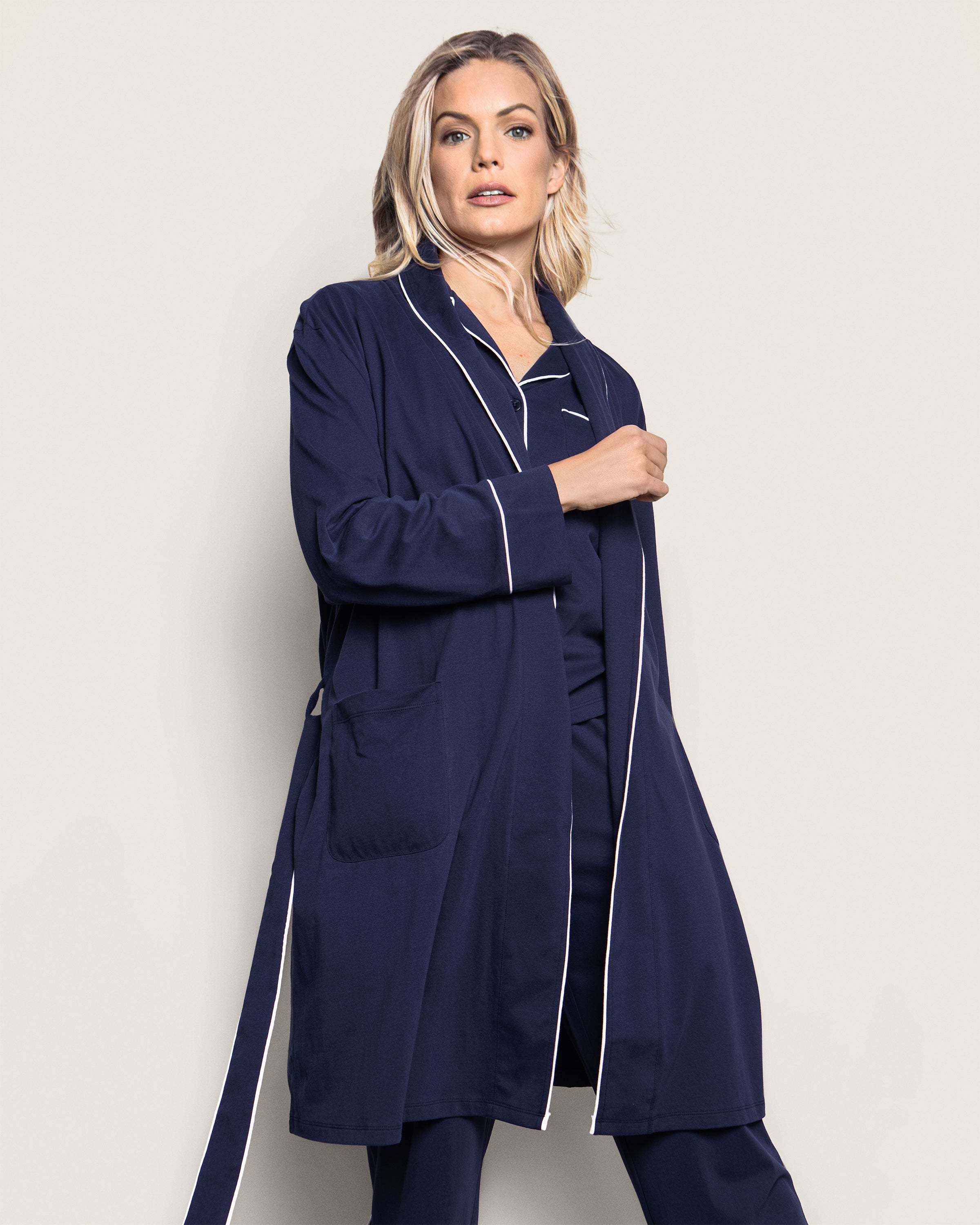 A person with light-colored hair stands against a light background, wearing the Womens Pima Robe in Navy by Petite Plume. This luxury robe, crafted from soft Peruvian Pima cotton, features white piping, a tied belt, and a large pocket. They are looking slightly off-camera.