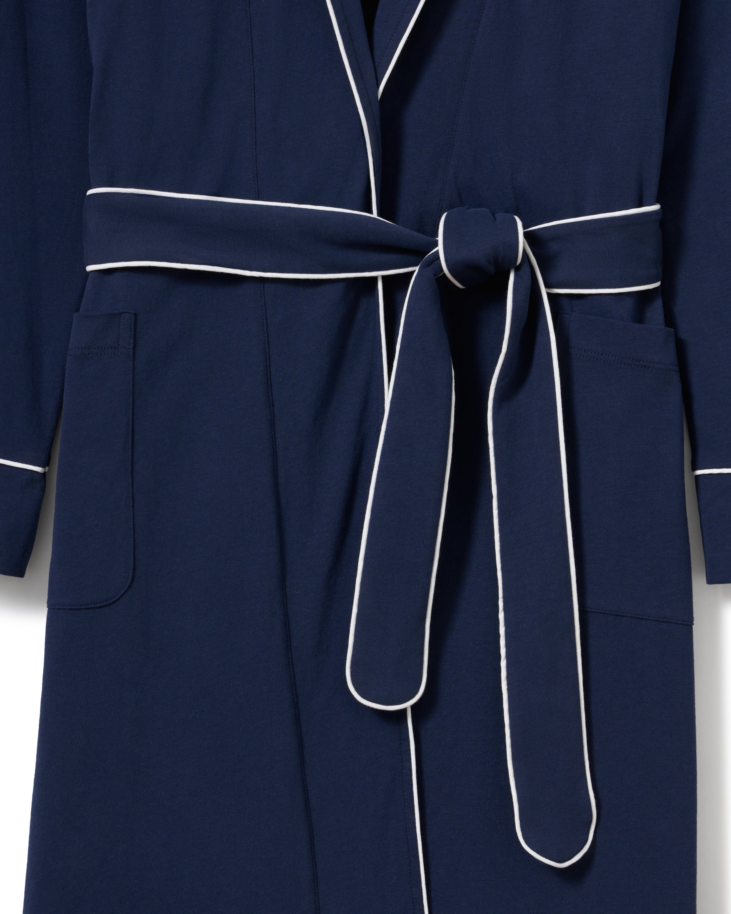 A person with long hair stands confidently in dark blue luxury pajamas made from yarn-dyed Peruvian Pima cotton, highlighted with white piping. They have the Womens Pima Robe in Navy by Petite Plume draped over their shoulders, hands in pockets, set against a light beige background.