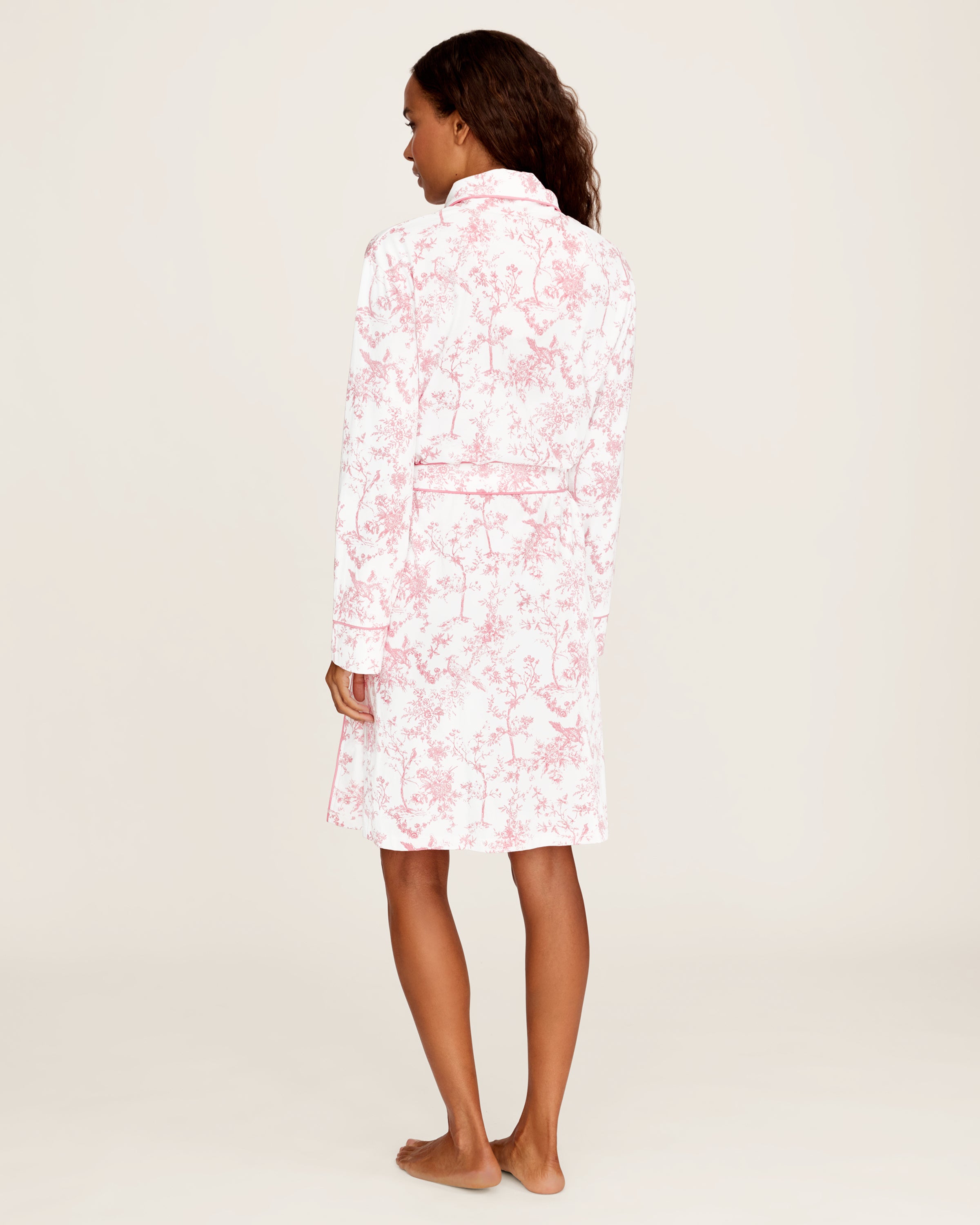 The Women's Pima Robe in Pink Timeless Toile features a white knee-length design with a pink floral toile pattern, long sleeves, a shawl collar, and a matching waist belt.