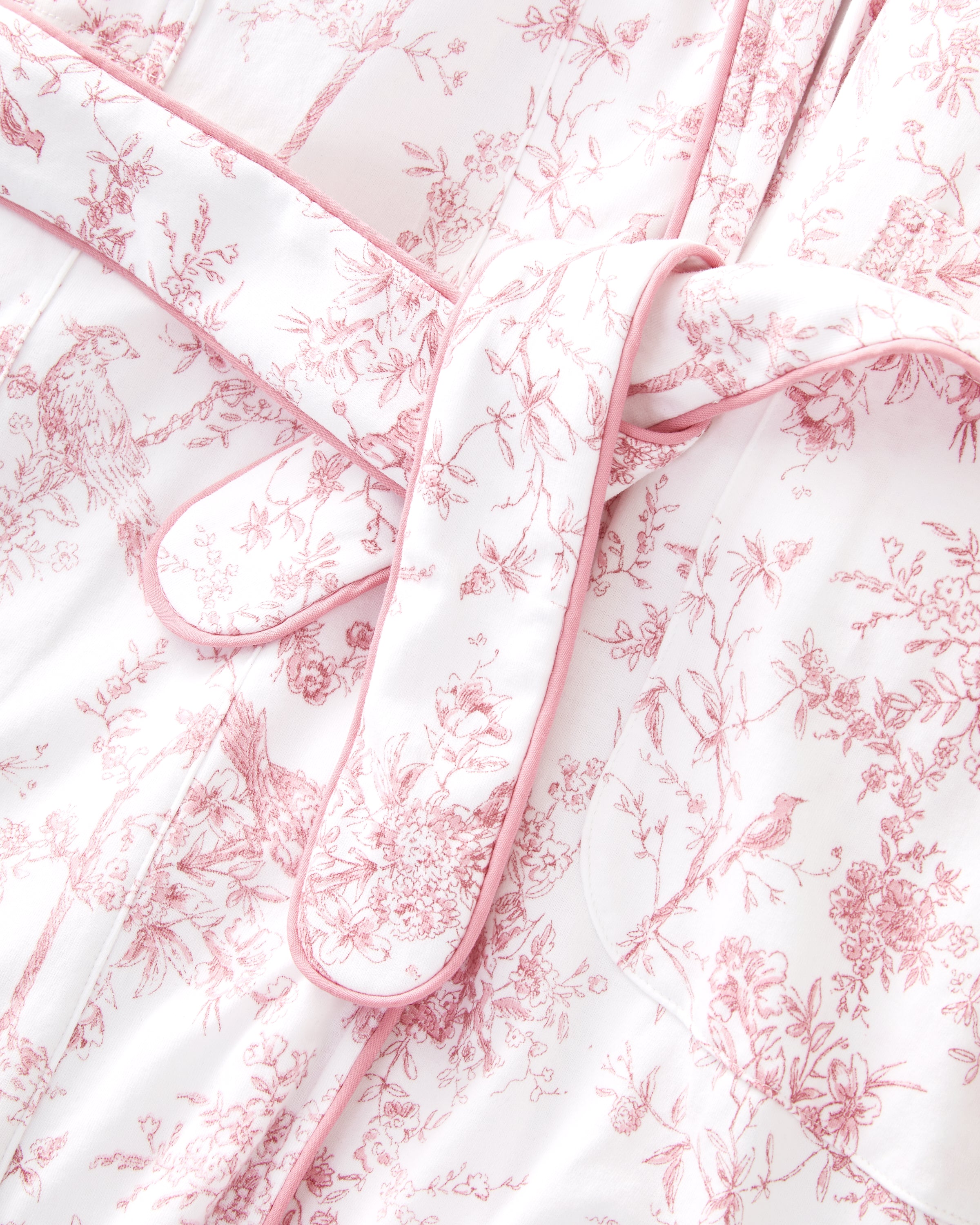 The Women's Pima Robe in Pink Timeless Toile features a white knee-length design with a pink floral toile pattern, long sleeves, a shawl collar, and a matching waist belt.
