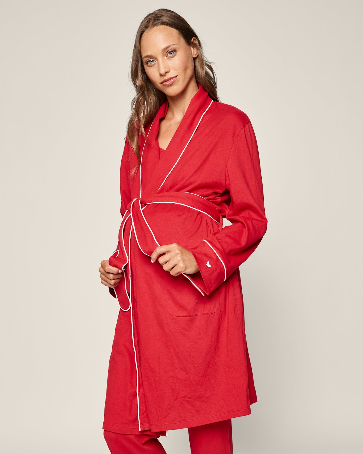 A person with long hair dons the Womens Pima Maternity Robe in Red by Petite Plume. Crafted from soft Peruvian Pima cotton, it boasts white trim, a V-neck, and wide sleeves. They stand against a plain background, holding the belt with both hands.
