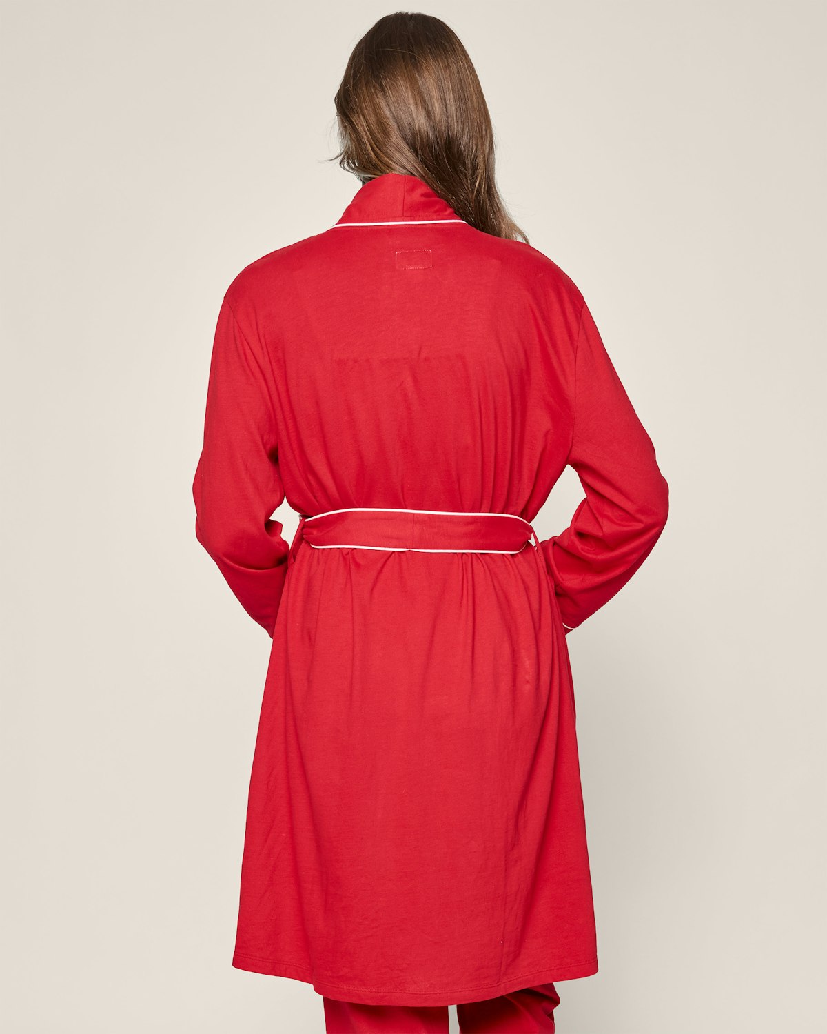 Facing away from the camera, a person is wrapped in Petite Plumes Womens Pima Maternity Robe in Red, featuring white piping and a matching fabric belt. Long brown hair cascades against the plain beige background. Crafted from soft Peruvian Pima cotton, this robe exudes elegance.