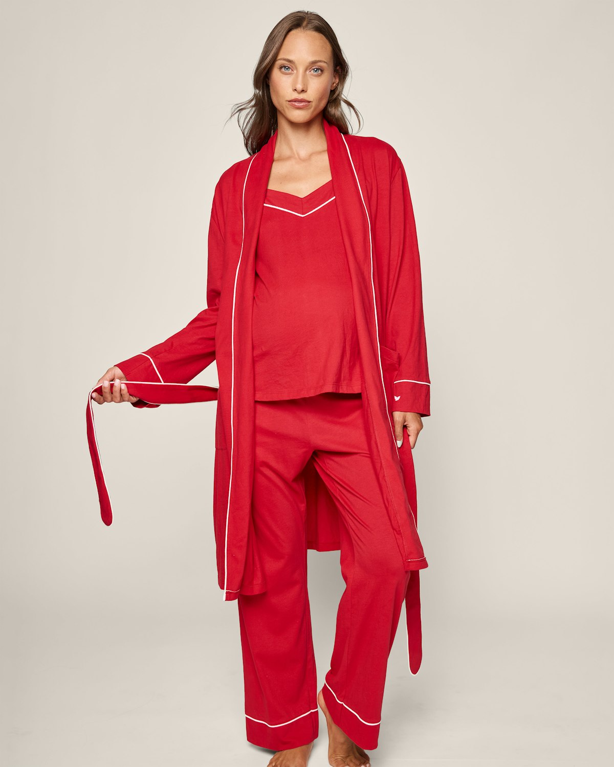 A woman in Petite Plumes Womens Pima Maternity Robe in Red, featuring white piping, poses against a plain light backdrop. She holds the robes belt in one hand, looking directly at the camera.