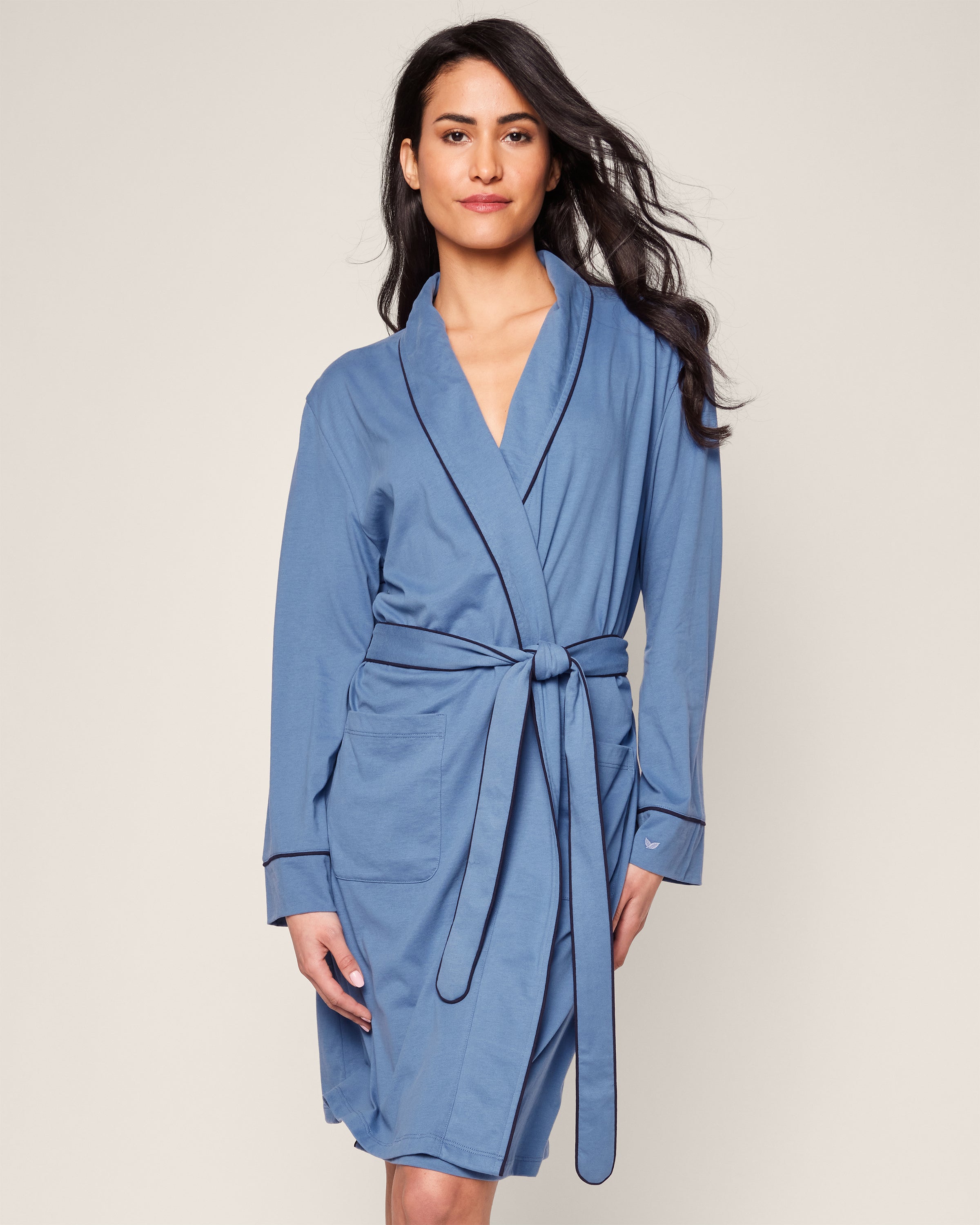 A woman showcases the Womens Pima Robe in Mystique Blue by Petite Plume against a plain backdrop. The robe, made from Pima cotton with dark edging and a sash belt, complements her long wavy hair and slight smile, embodying luxury sleepwear elegance.
