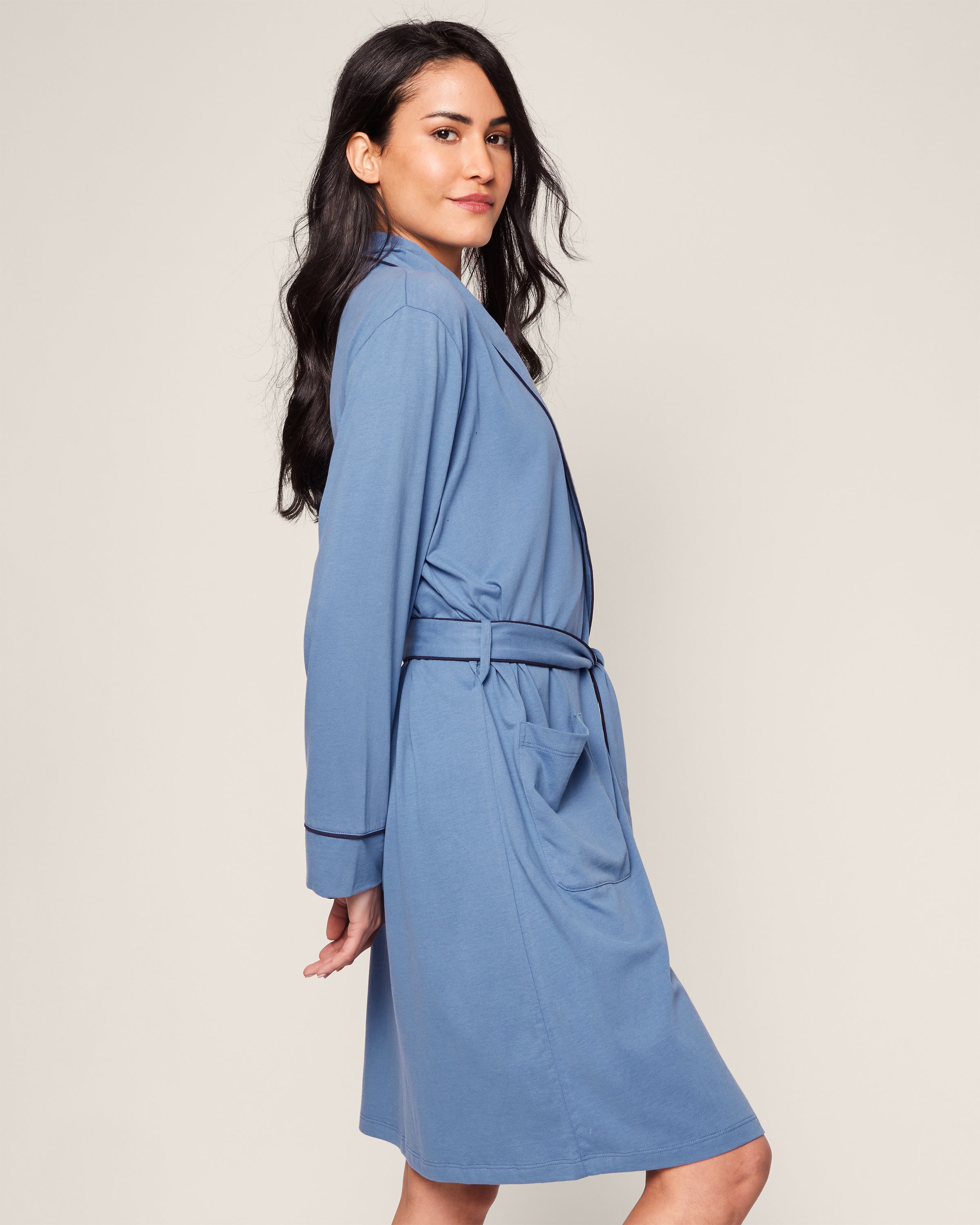 A woman with long dark hair wears Petite Plumes Womens Pima Robe in Mystique Blue. Crafted from Pima cotton, she stands sideways against a neutral background, showcasing the robes long sleeves and knee-length hem, epitomizing luxury sleepwear.