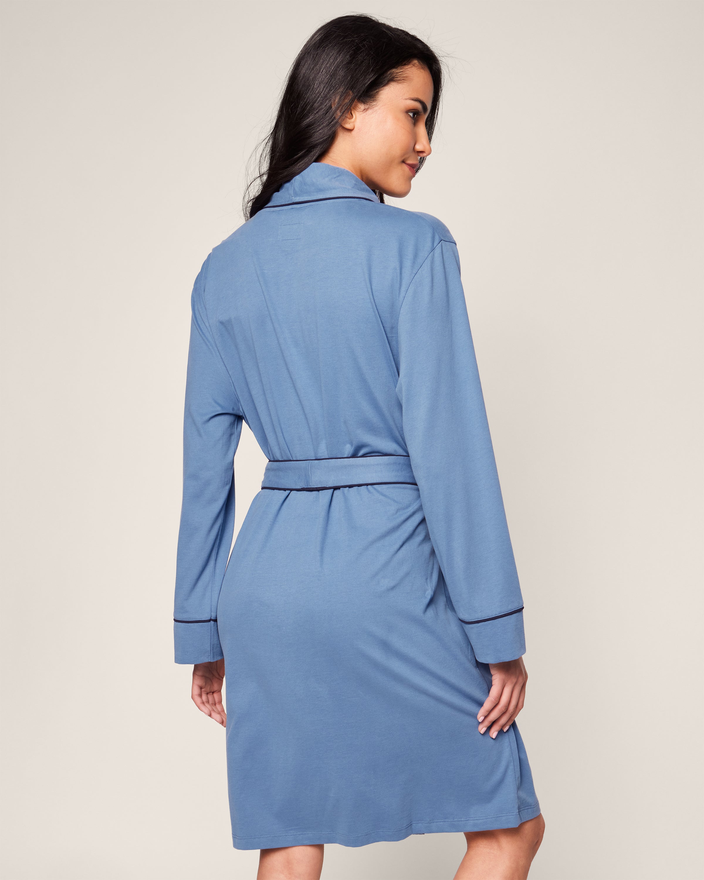 A woman with long dark hair wears an elegant Womens Pima Robe in Mystique Blue by Petite Plume, made of Pima cotton, featuring a belt and dark piping on the collar and sleeves. She smiles gently over her shoulder against a plain backdrop.