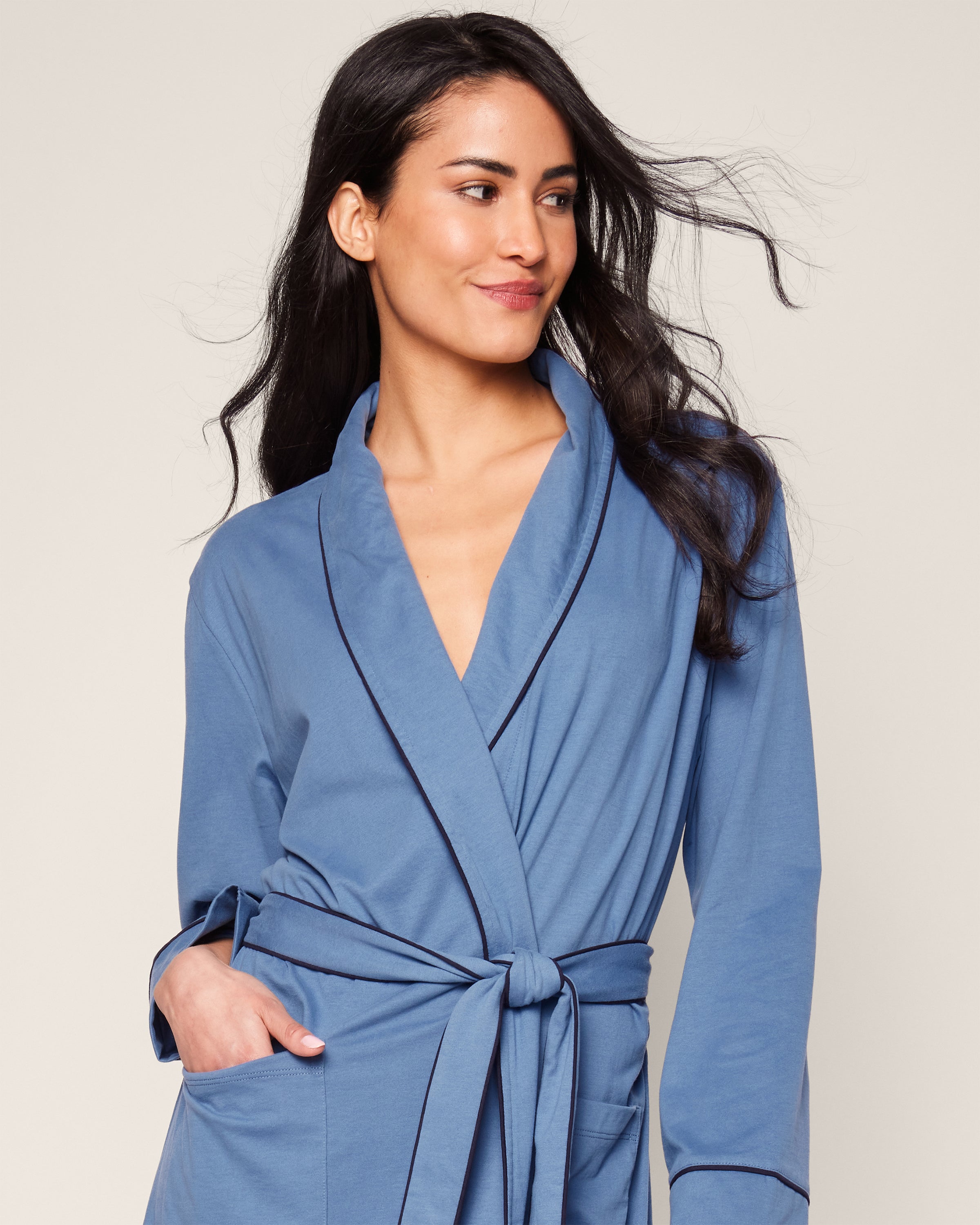 With long, dark hair, the person wears the Womens Pima Robe in Mystique Blue by Petite Plume. Made from luxurious cotton, it features a sash tie, pockets, and dark trim. They glance sideways with a gentle smile against a plain, light backdrop.