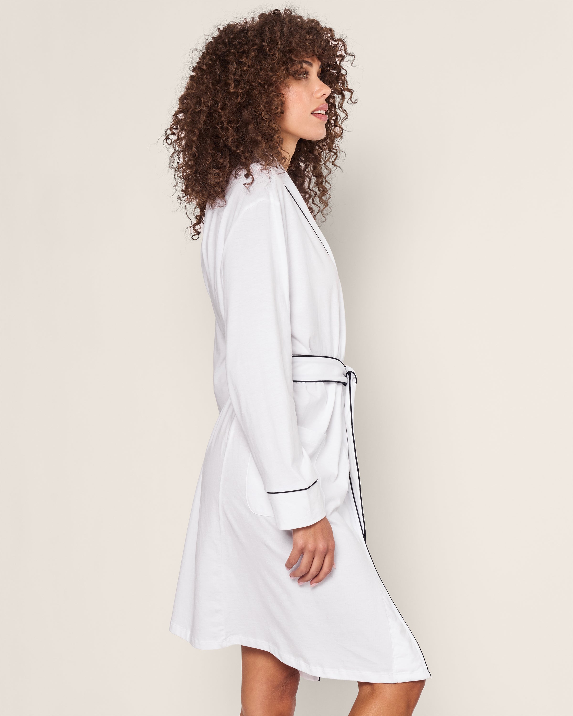 A person with curly hair is elegantly dressed in a Womens Pima Robe in White with Navy Piping by Petite Plume, standing in profile against a plain background.