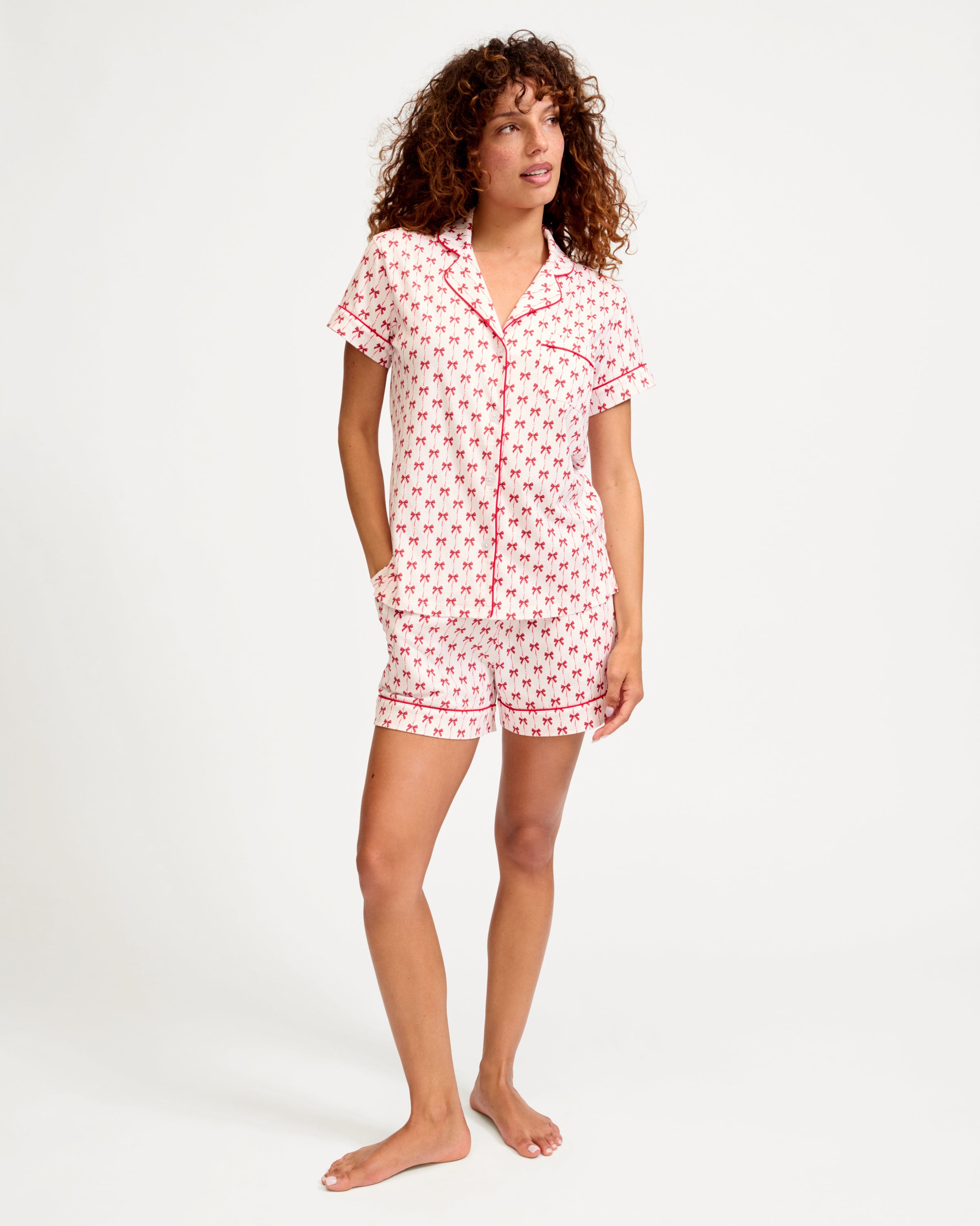 The Women's Pima Short Set in Blissful Bows features a short-sleeve white pajama top and shorts, adorned with a red palm tree pattern and red piping on the edges, collar, pocket, and cuffs.