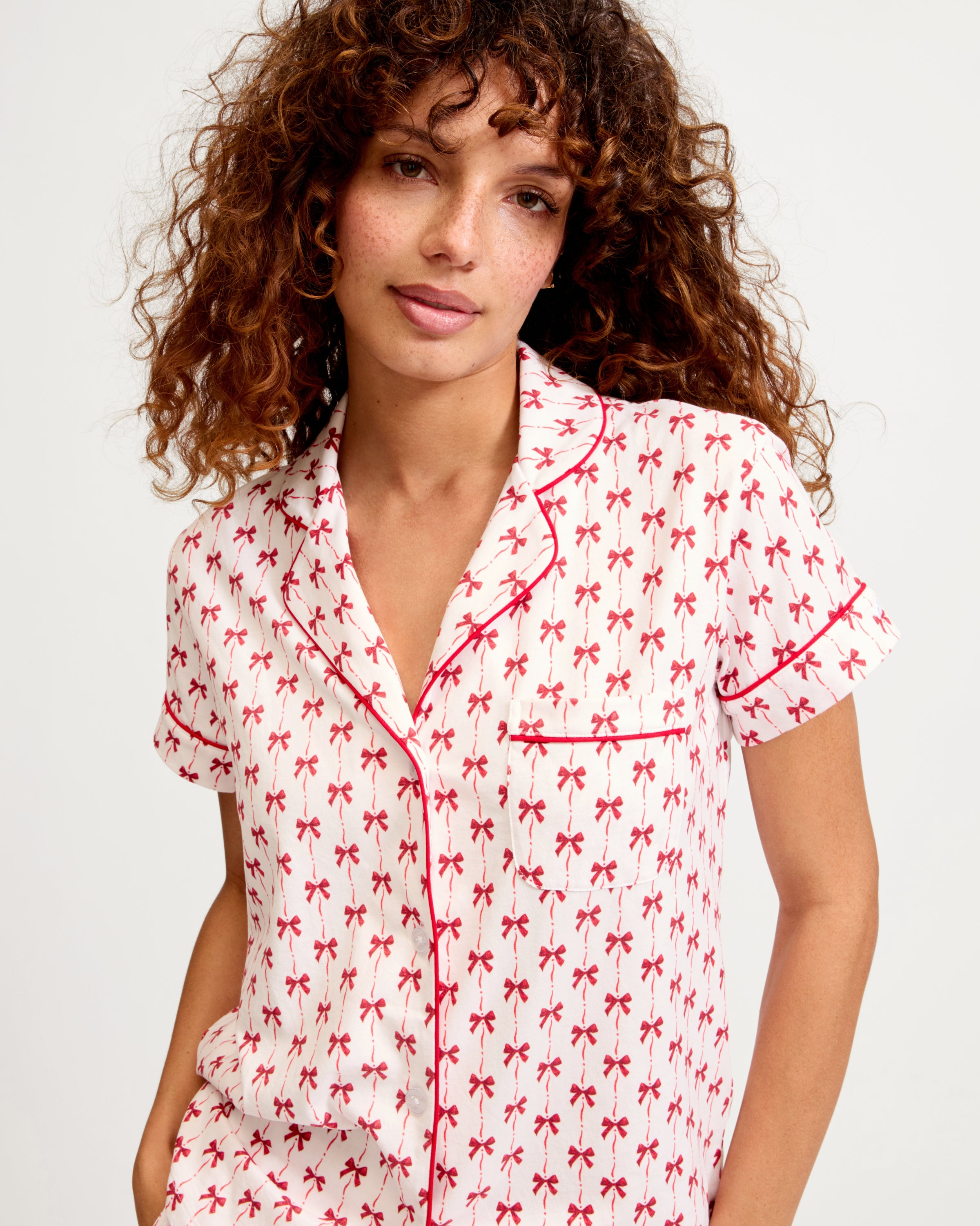 The Women's Pima Short Set in Blissful Bows features a short-sleeve white pajama top and shorts, adorned with a red palm tree pattern and red piping on the edges, collar, pocket, and cuffs.