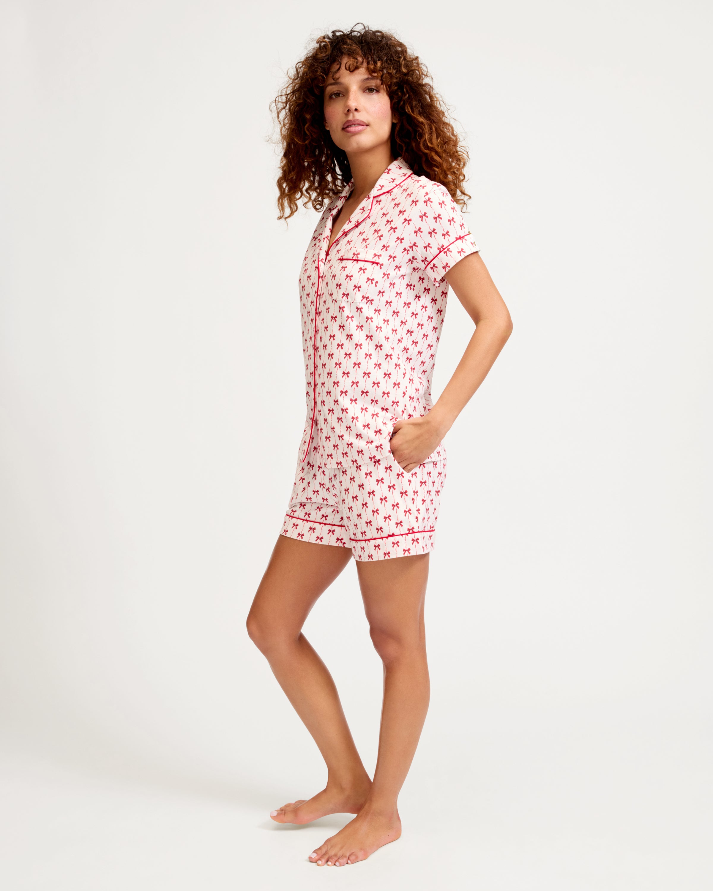 The Women's Pima Short Set in Blissful Bows features a short-sleeve white pajama top and shorts, adorned with a red palm tree pattern and red piping on the edges, collar, pocket, and cuffs.