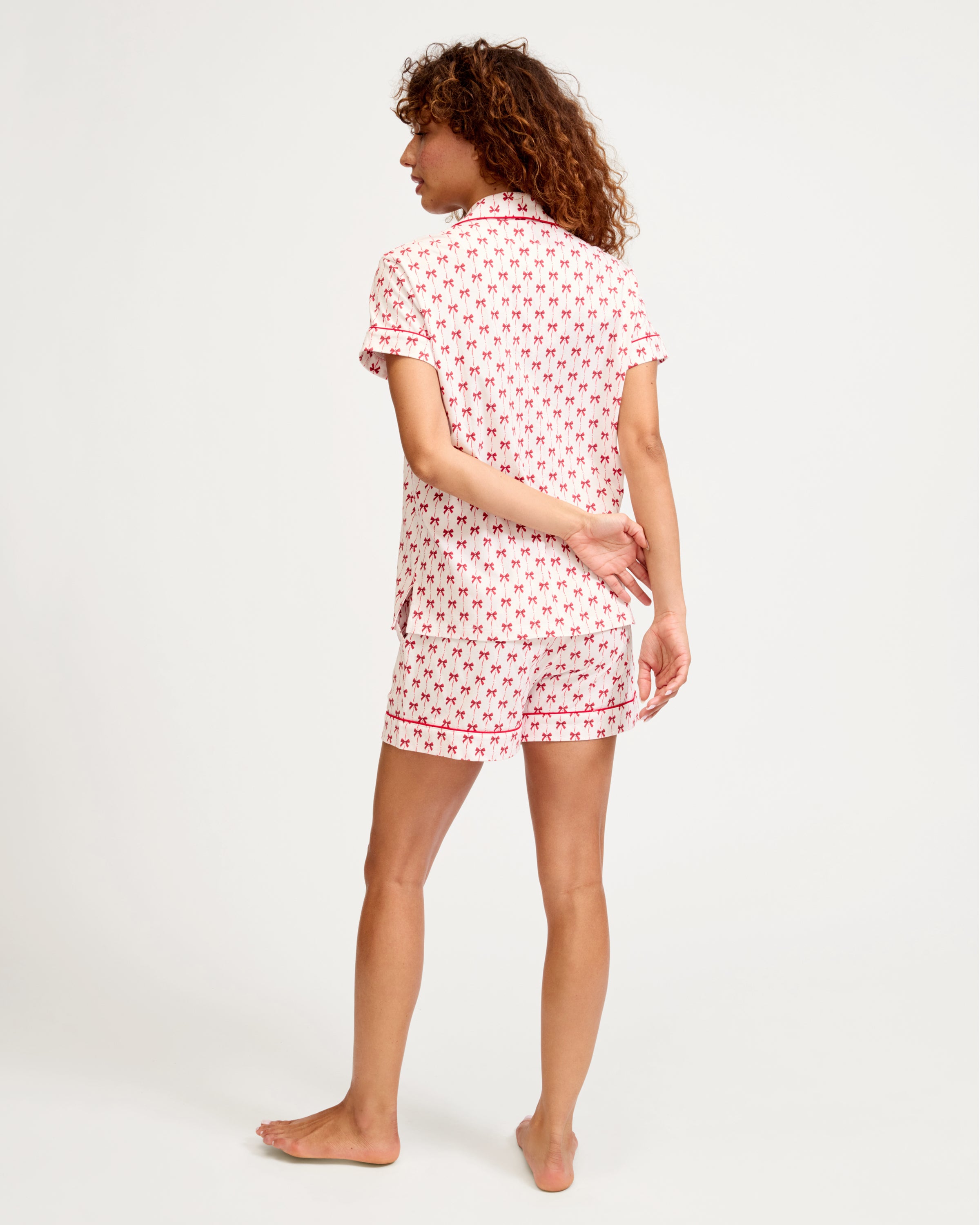 The Women's Pima Short Set in Blissful Bows features a short-sleeve white pajama top and shorts, adorned with a red palm tree pattern and red piping on the edges, collar, pocket, and cuffs.