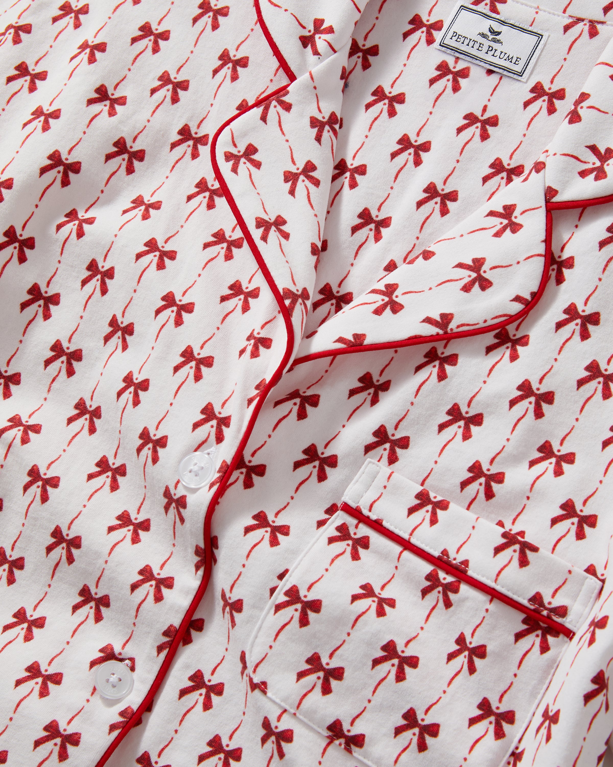 The Women's Pima Short Set in Blissful Bows features a short-sleeve white pajama top and shorts, adorned with a red palm tree pattern and red piping on the edges, collar, pocket, and cuffs.