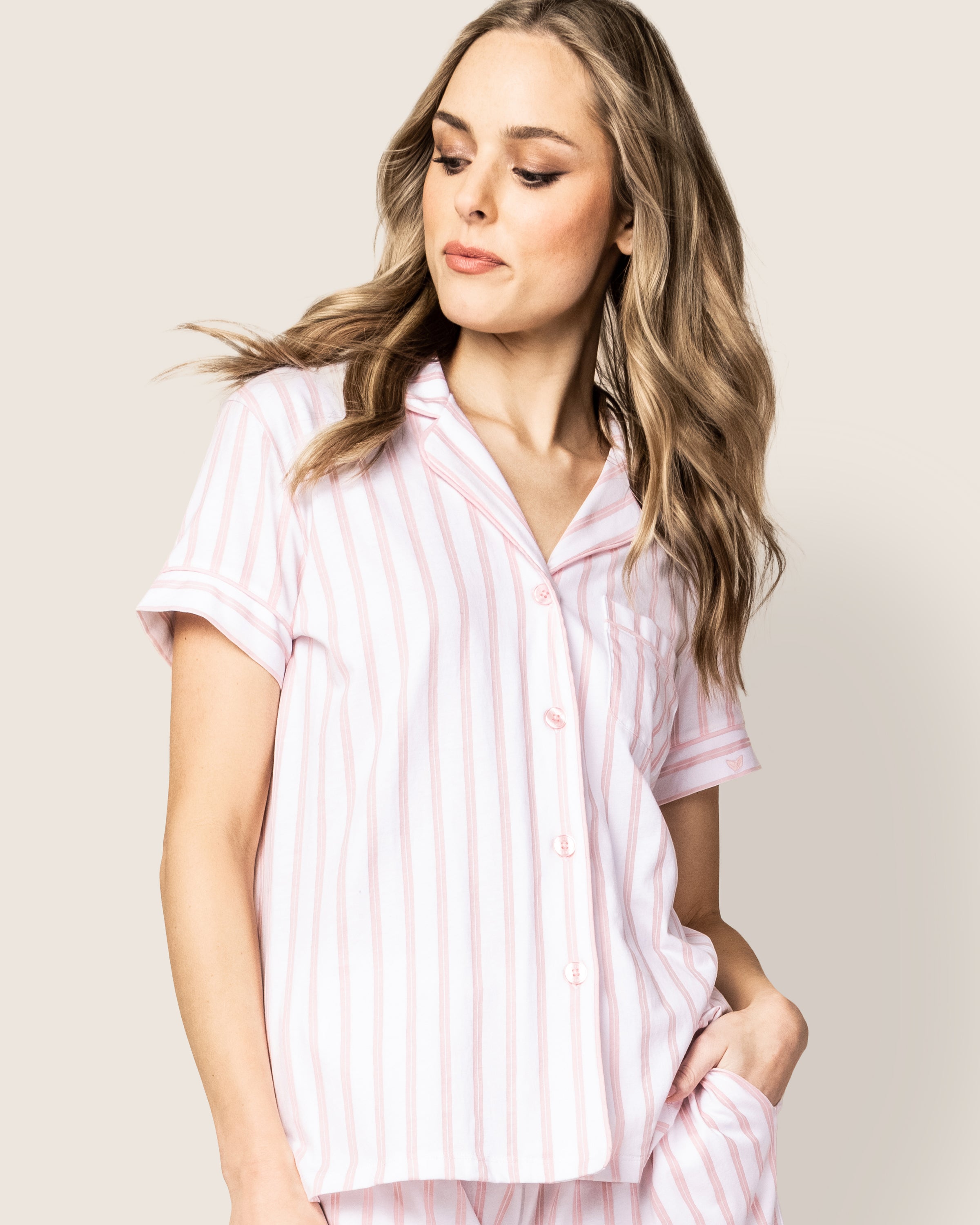 A person with long hair wears Petite Plumes Womens Pima Pajama Short Set in Pink Stripe, featuring a short-sleeved button-up shirt and matching pants. They stand against a plain background, gazing to the side with one hand in their pocket, exuding elegance.