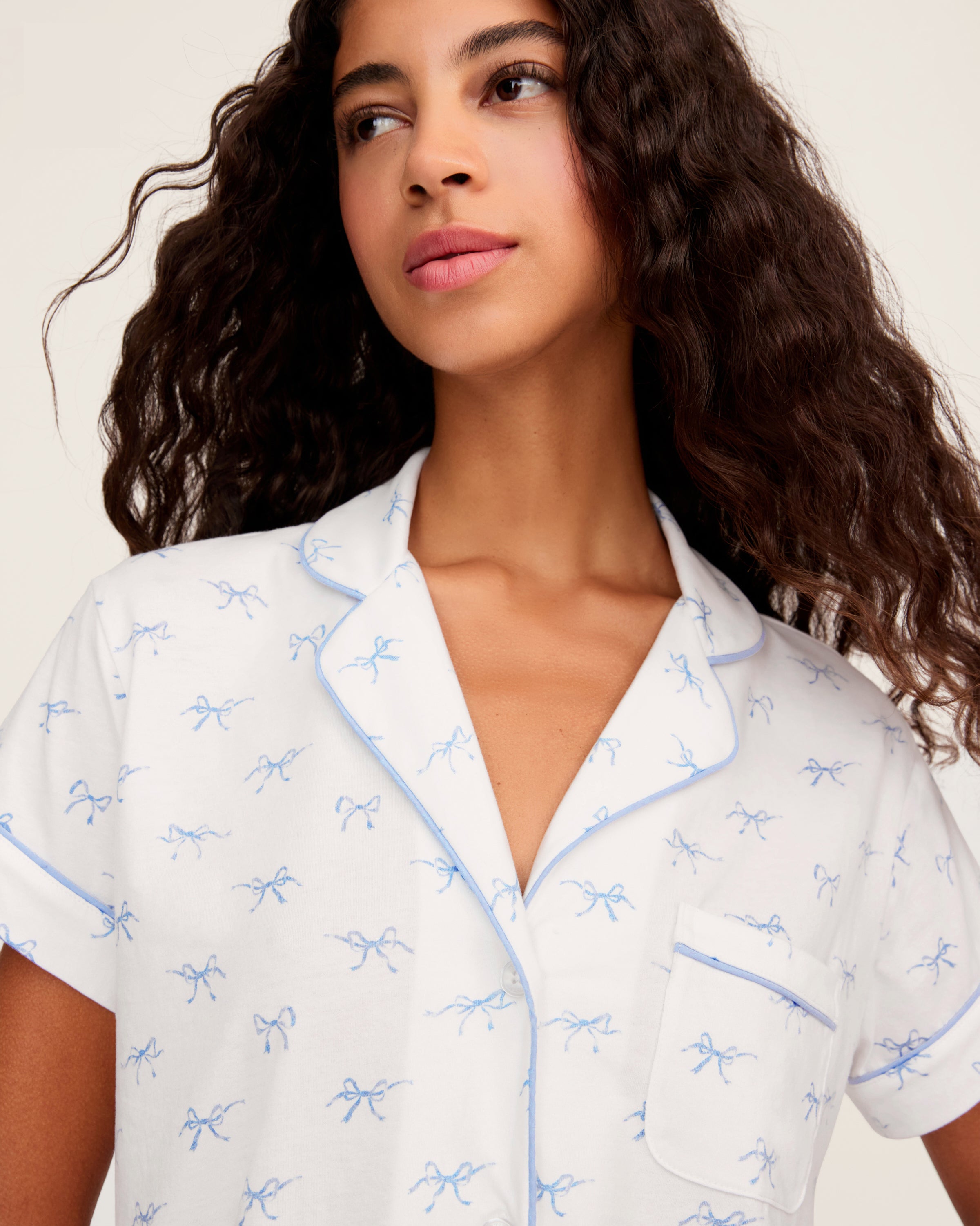 Women's Pima Short Sleeve Short Set in Something Blue Bows features a collared button-up top with chest pocket and matching elastic-waist shorts, both adorned with all-over blue bow print and contrast piping.